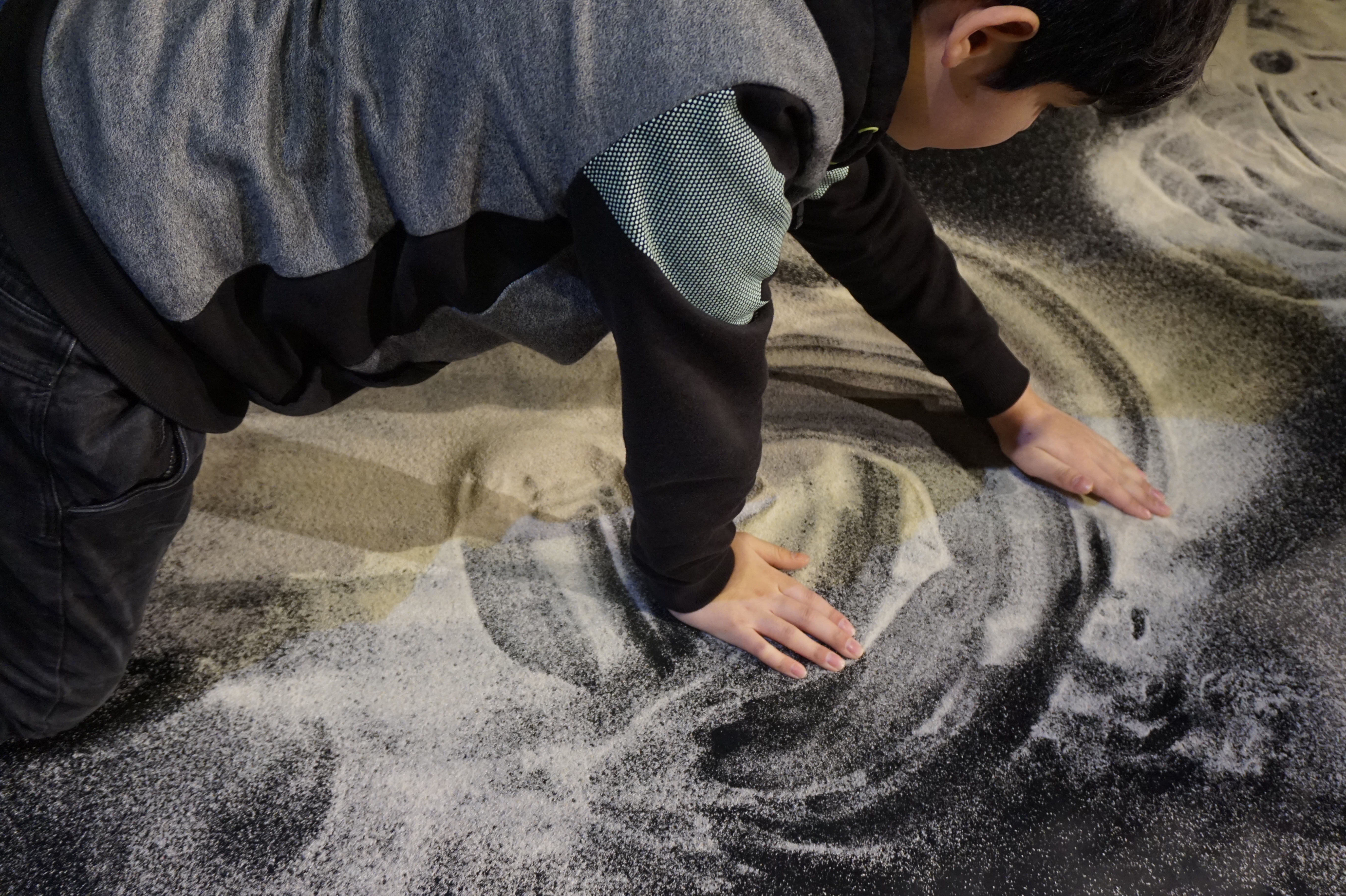 Ein KInd wischt mit den Händen ein Muster in Sand auf dem Boden