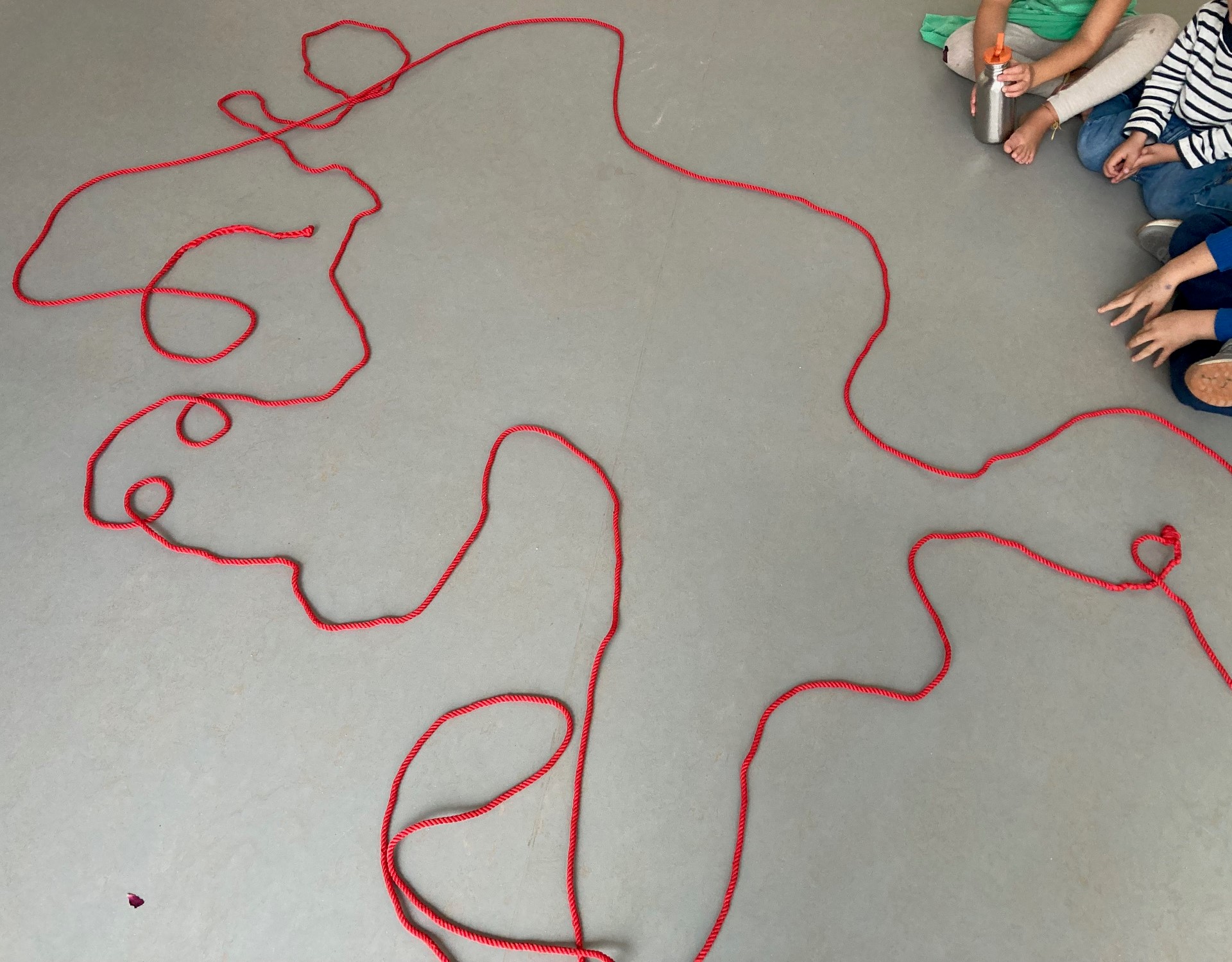 auf einem grauen Boden liegt ein großes rotes Seil, das durch seine vielen Rundungen,Schlingen und Kurven wie ein kleines Monsterwesen aussieht, am Rand sieht man Hände und knie von Kindern die darum sitzen