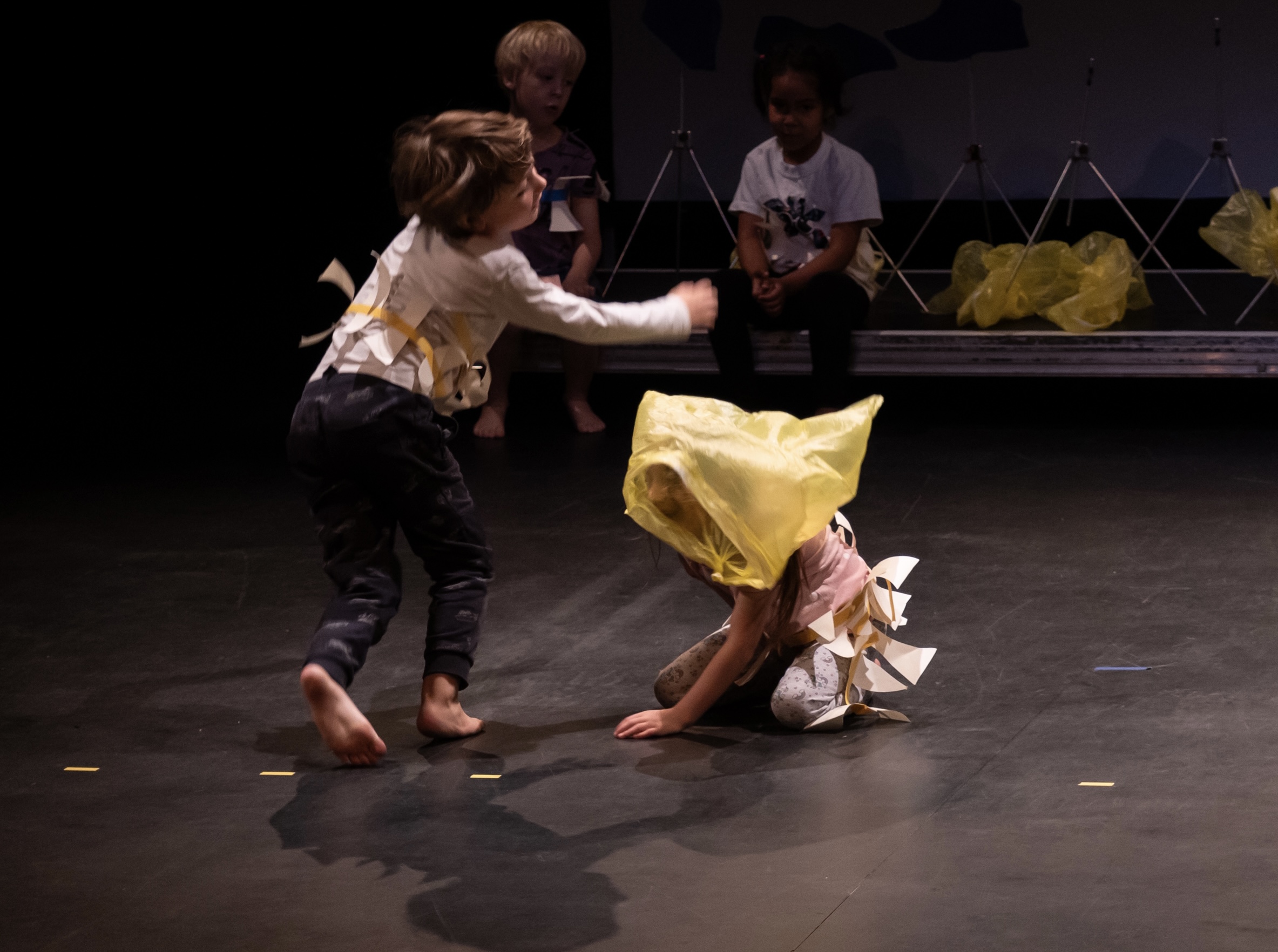 zwei kinder auf der Bühne mit weißem oberteil und einer gelben Tüte in der Hand bewegen sich. Ein kind hält die Arme auseinander, das andere ist auf allen vieren am Boden