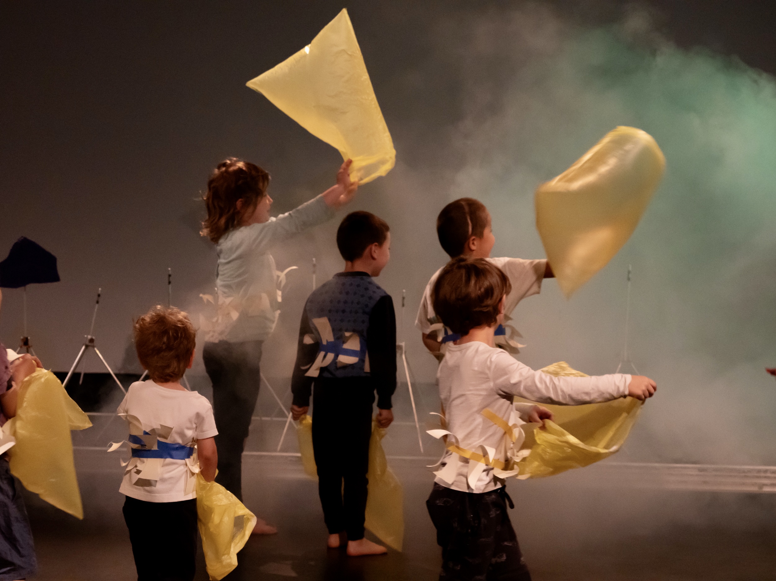 mehrere Kinder fangen Luft in gelbe Plastiktüten ein. Sie speieln in einem Bühnenraum mit gelb grünen Licht und im neben
