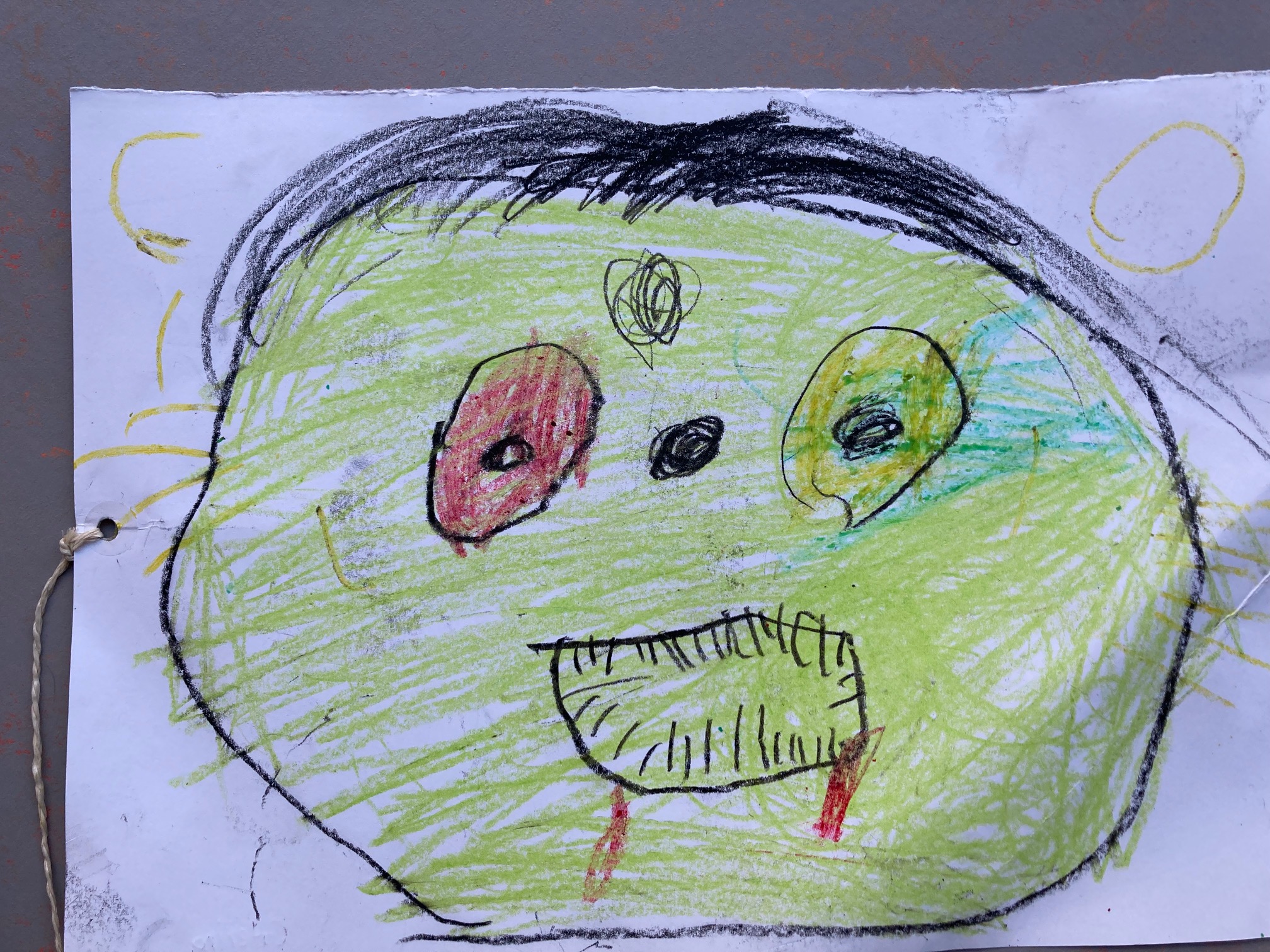 auf einem weißen Blatt sieht man ein grünes Gesiht mit einem roten und einem grünen Auge. Der Mund ist als rundliche Öffnung mit vielen kleinen Strichen, die Zähne andeuten, zu sehen. Die Nase ist ein schwarzer Punkt. es gibt außerdem schwarze kurze Haare