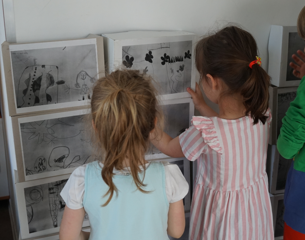 zwei Mädchen haben an der Wand eine mauer aus kartons gestapelt. Auf der Mauer sind schwarz-weiße Kinderzeichnungen zu sehen. Die Mädchen sind von hinten zu sehen und tragen einen Pferdeschwanz.