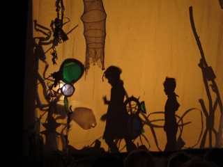 Schattenprojektion aus Silhouetten und Material auf gelb angeleuchtetem Hintergrund. Die seitlichen Schatten von zwei Kindern sind zu sehen sowie filigrane Formen, die an Türme erinnern. Oben zeichnet sich der Schatten einer Fischreuse ab.