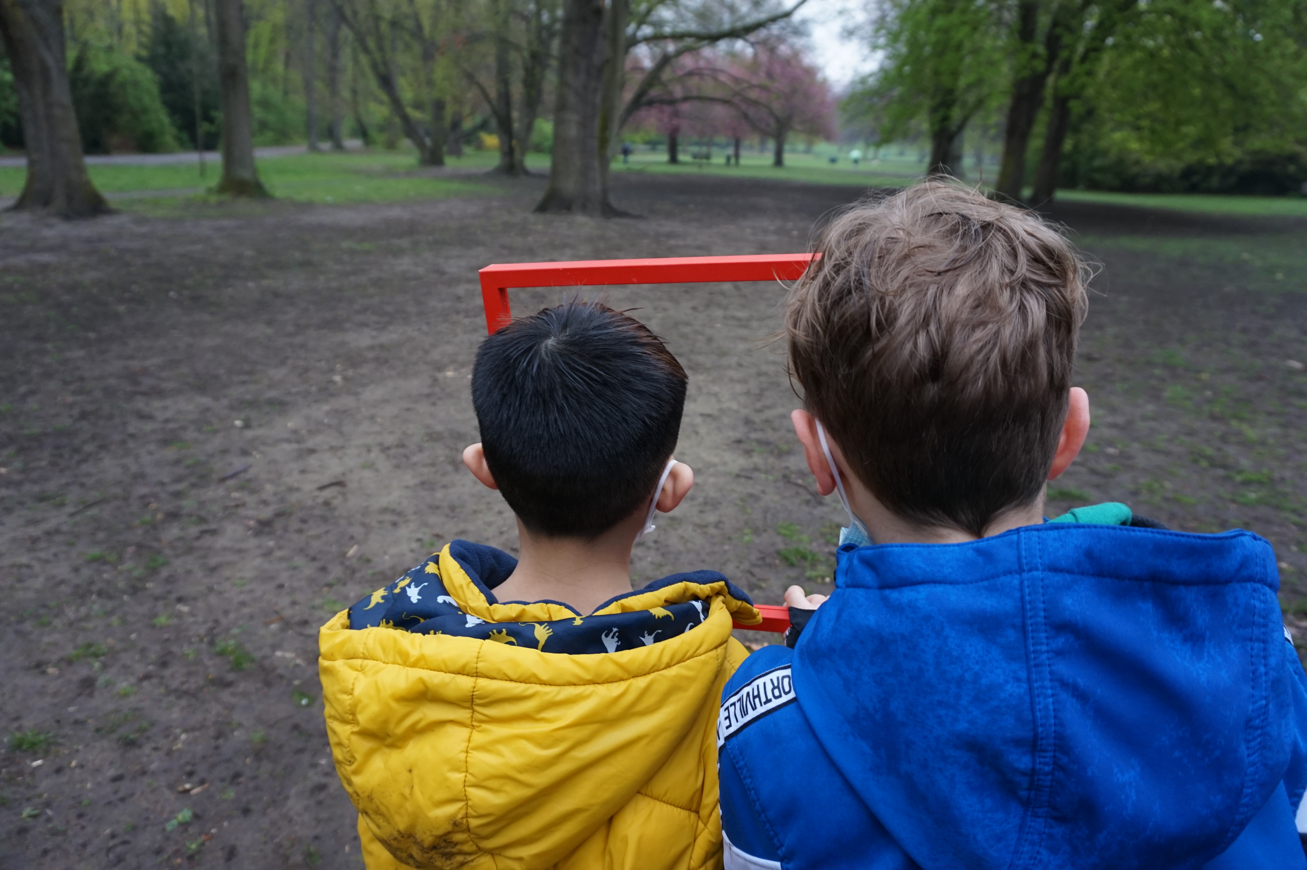 Zwei Kinder im Alter von etwa sieben oder acht Jahren sind von hinten zu sehen, wie sie im Park durch einen roten Rahmen schauen.