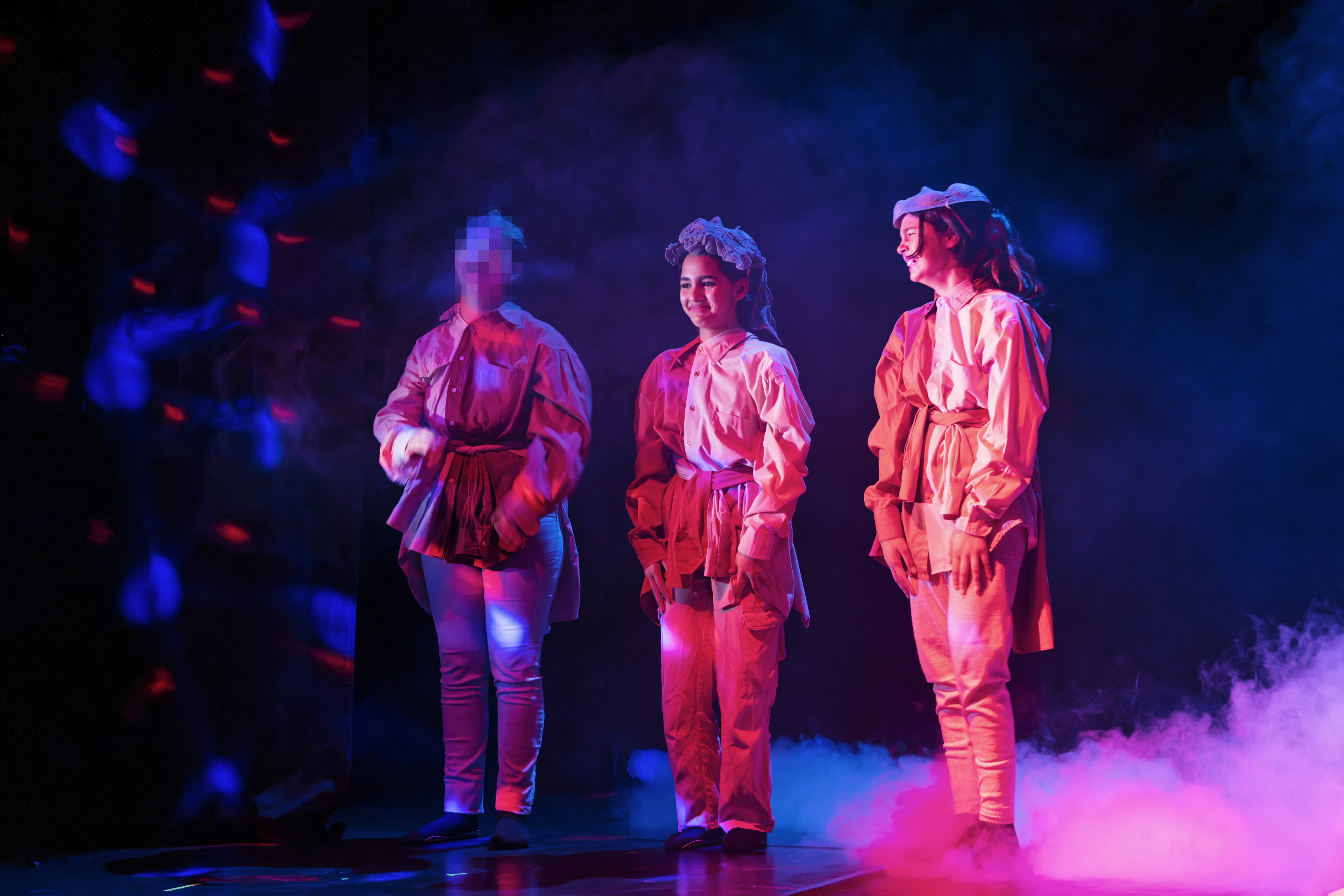 drei Mädchen stehen in einem rötlichen Show-Licht und lachen - von links sieht man Nebel, der sich um die Füße der Mädchen ausbreitet. Sie tragen Herrenhemden übereinander und verknotet