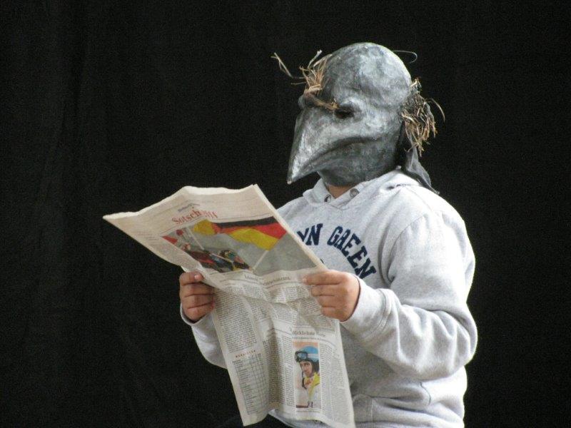 Ein Spieler steht mit einer grauen Vogelmaske mit spitzem Schnabel zeitungslesend im Bild