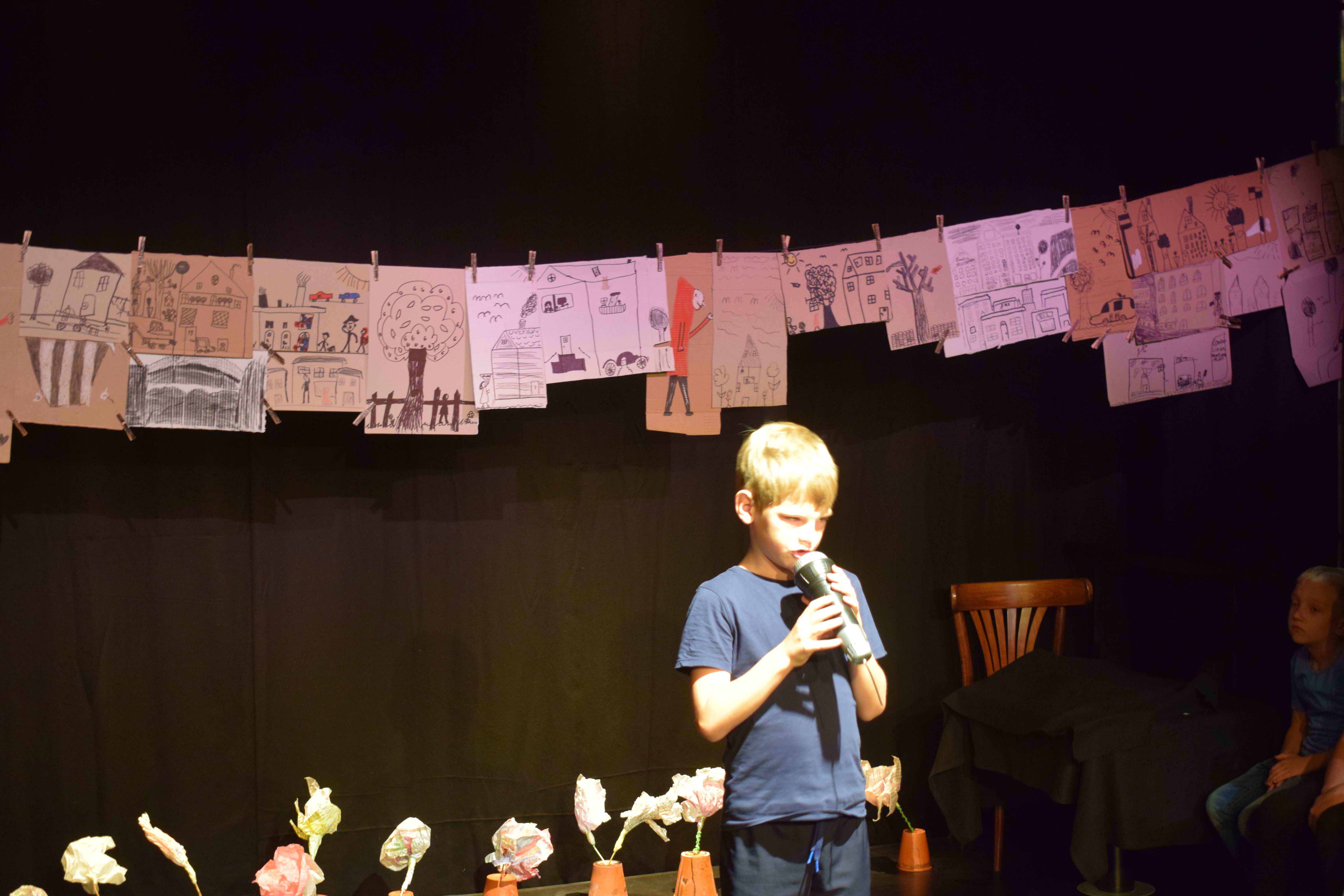 Ein etwa siebenjähriger Junge steht auf der Bühne mit einem Mikrophon in beden Händen. Über ihm sind Kinderzeichnungen an einer Schnur aufgereiht. Hinter ihm stehen im unteren Bildbereich umgedrehte Blumentöpfe, in denen gebastelte Papierblumen stecken. Rechts zeichnen sich im Dunkel zwei Kinder an, die den Jungen anschauen.