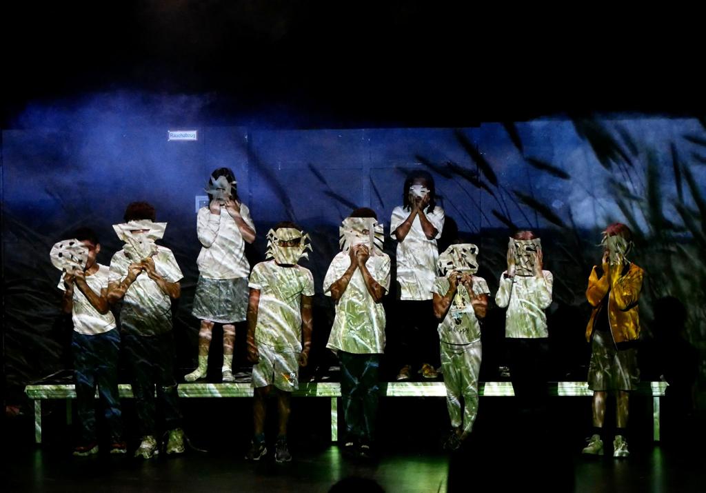 Auf einem Podest stehen Kinder mit Masken vor blauem Hintergrund mit Grasschatten. Die Kleidung der Kinder ist mit grünen Strichen angeleuchtet