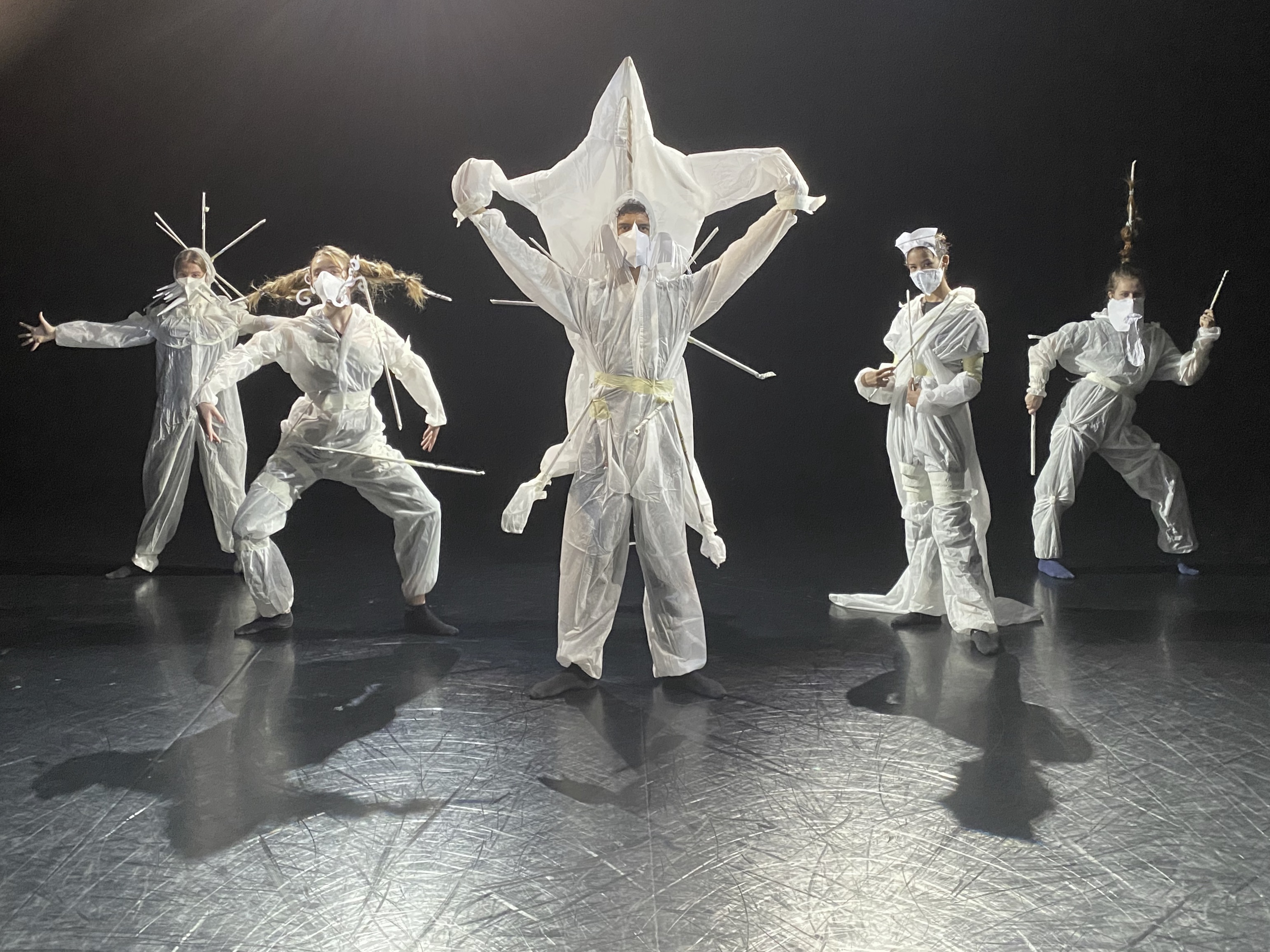 Fünf in weiße Schutzanzüge gekleidete Personen tragen futuristisch wirkende Masken aus Papier und Stäben
