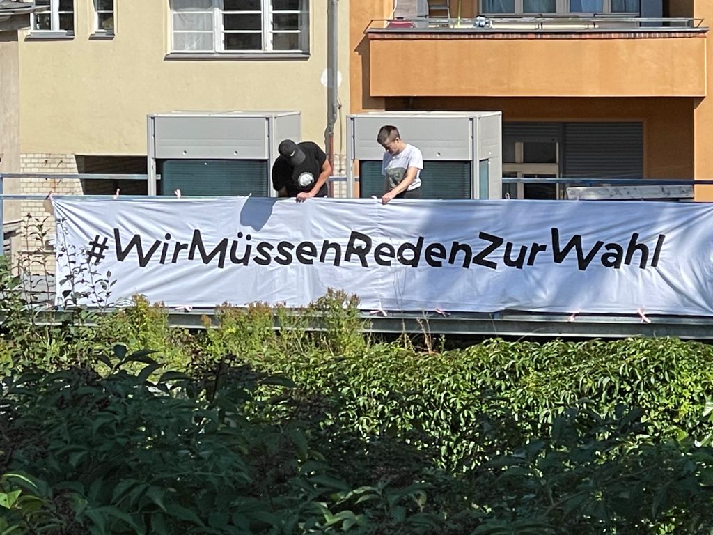 Two staff members of the Schaubude attaching a flag on the roof of the theatre that says #WirMüssenRedenZurWahl (We have to talk about the election)