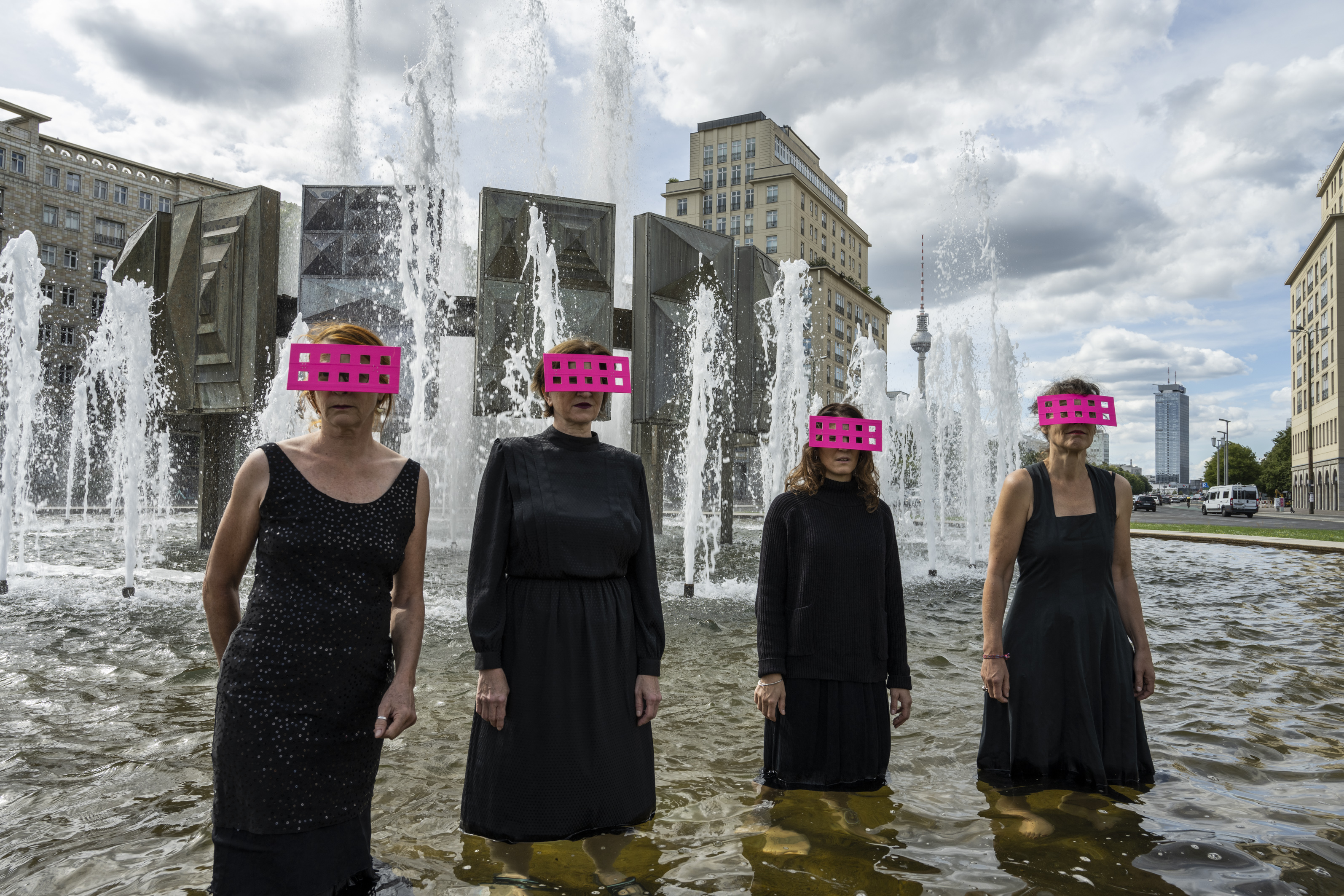 Vier Frauen stehen in dunklen Kleidern mit rosafarbenen, durchlöcherten Brettern vor den Augen in einem Brunnen in der Nähe vom Alexanderplatz.