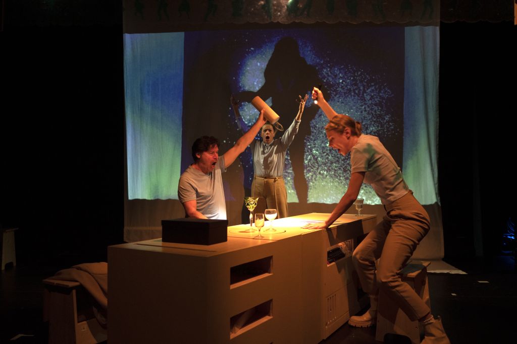 Zwei Schauspieler, ein Mann und eine Frauen sitzen, halb erhoben, einander gegenüber an einem kastenartigen Tisch und schwenken Gegenstände in der Hand. Im Hintergrund die Schattenprojektion einer Figur.
