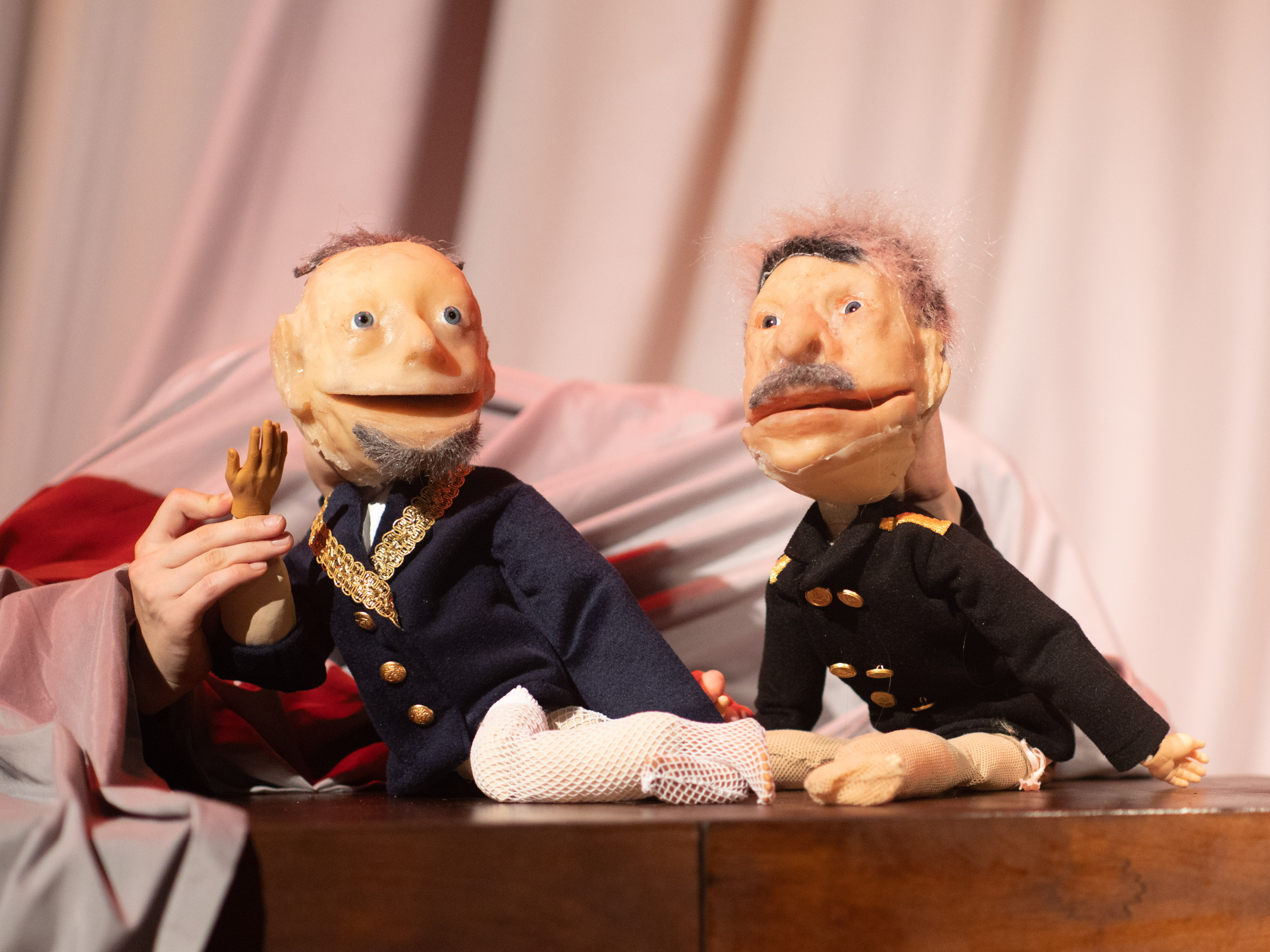 The puppets of two uniformed old men sit on a piece of wooden furniture. The two wear different beards and have gray hair. One of them has a bandage on his leg.