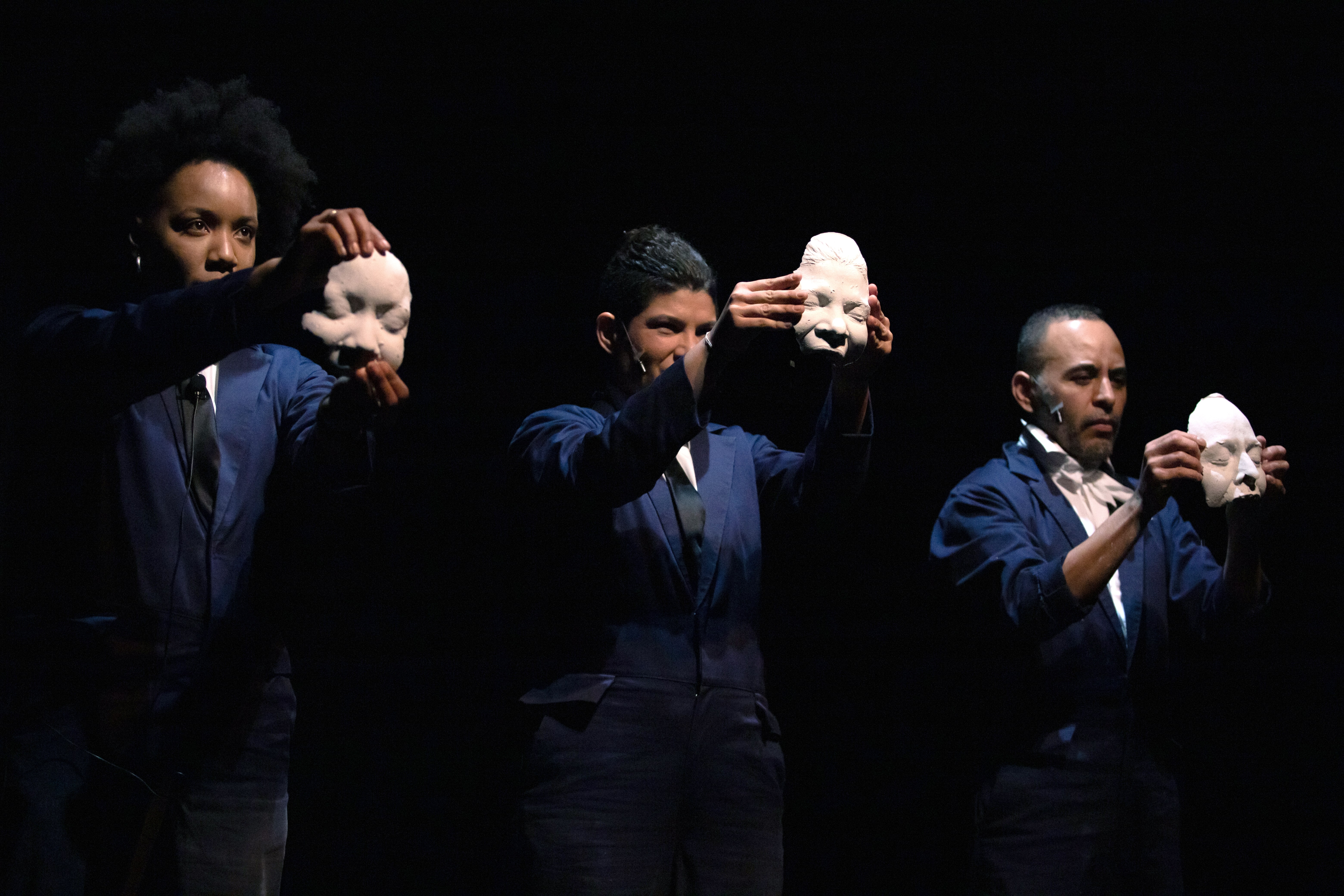 Drei Performende, zwei Frauen und ein Mann, stehen in identischen blauen Overalls auf der Bühne und halten in ihren Armen Gipsabdrücke ihrer Gesichter in die Höhe.