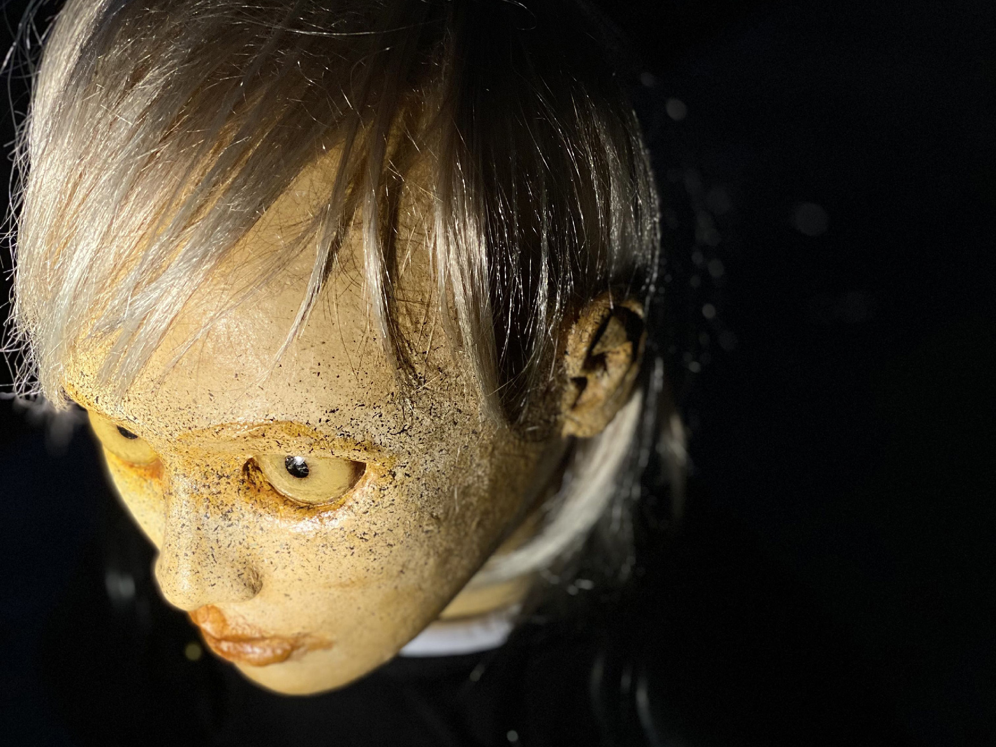 A doll with white hair and big yellow eyes looks rigidly to the left. The camera position is above the doll. Only the doll's head is visible, the background is dark.