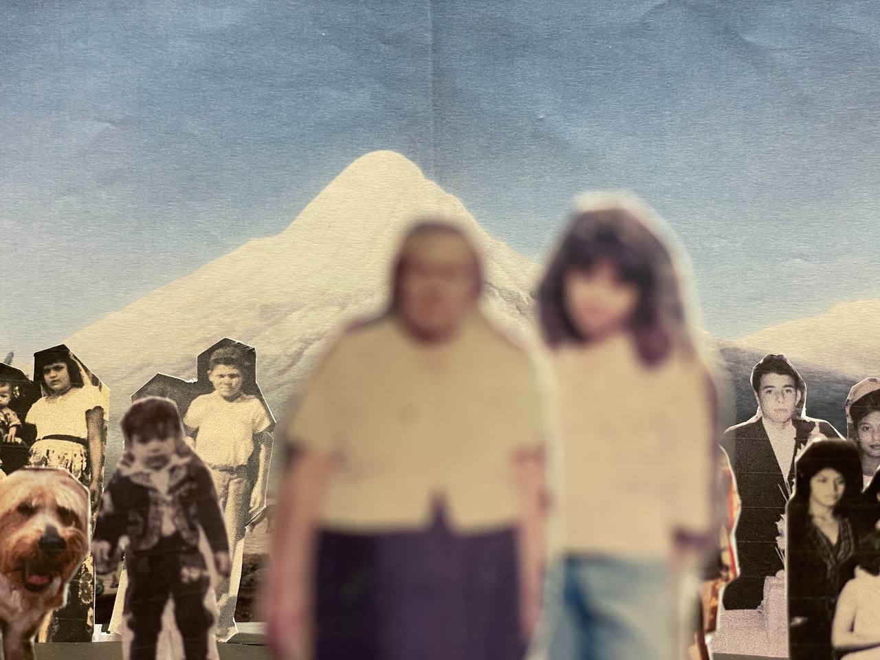 Das Bild zeigt eine Papiercollage, auf der ausgeschnittene Fotografien von acht Personen verschiedenen Alters und einem Hund zu sehen sind. Die zwei Personen im Vordergrund sind unscharf. Den Hintergrund bildet ein schneebedeckter Berg.
