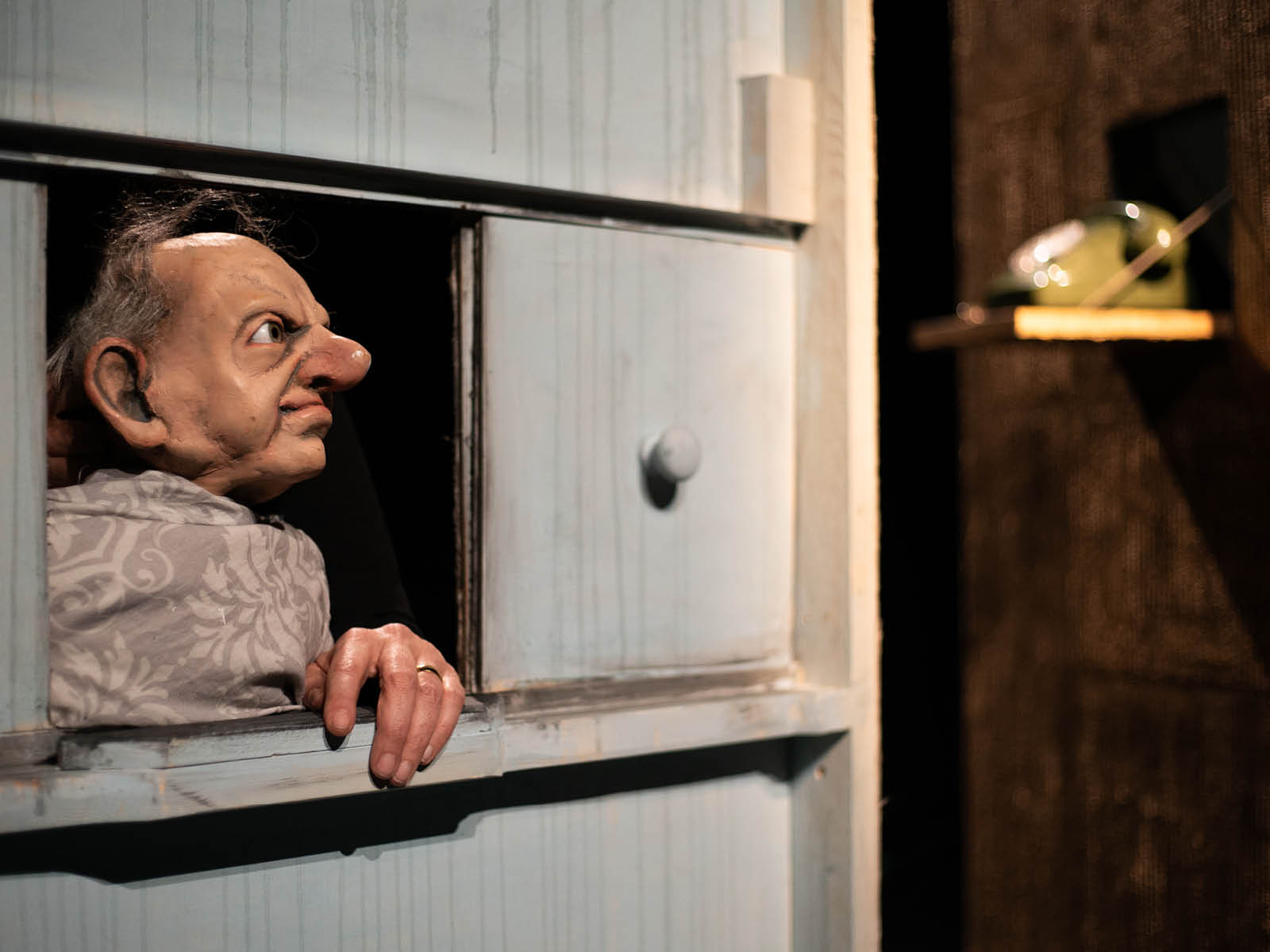 The doll of an old man with a half bald head looks out of a window. An old green telephone can be seen in the background.