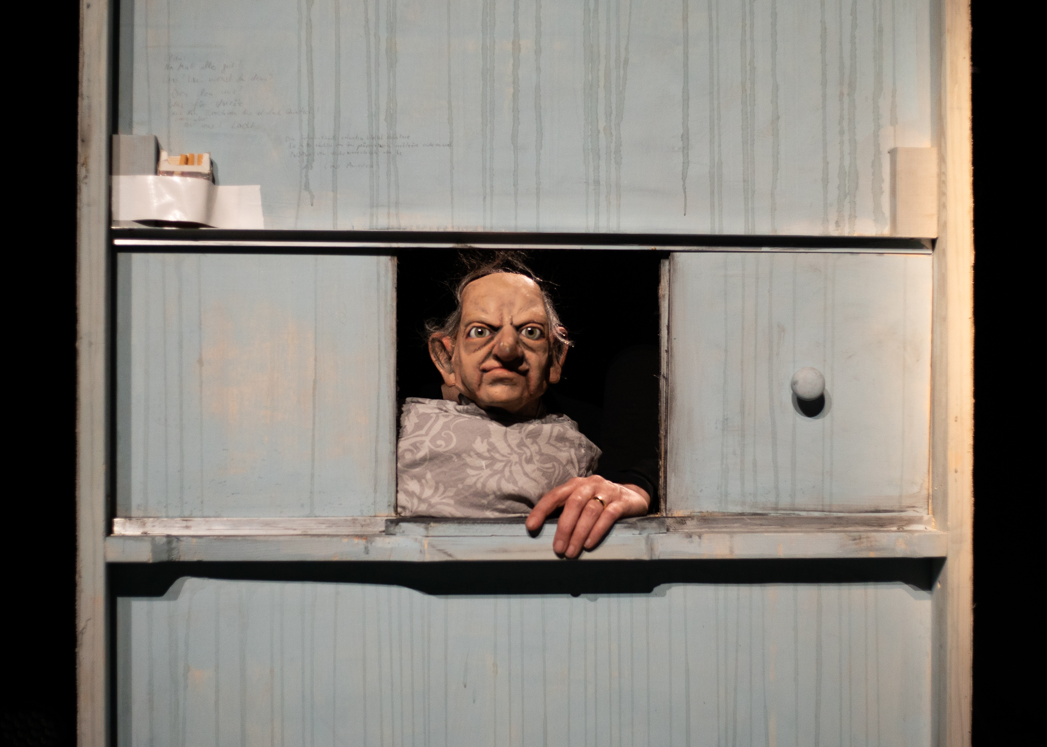 Die Puppe eines alten Mannes mit Halbglatze schaut aus dem Fenster Richtung Betrachter. Er stützt sich mit einer Hand auf dem Fensterbrett ab.
