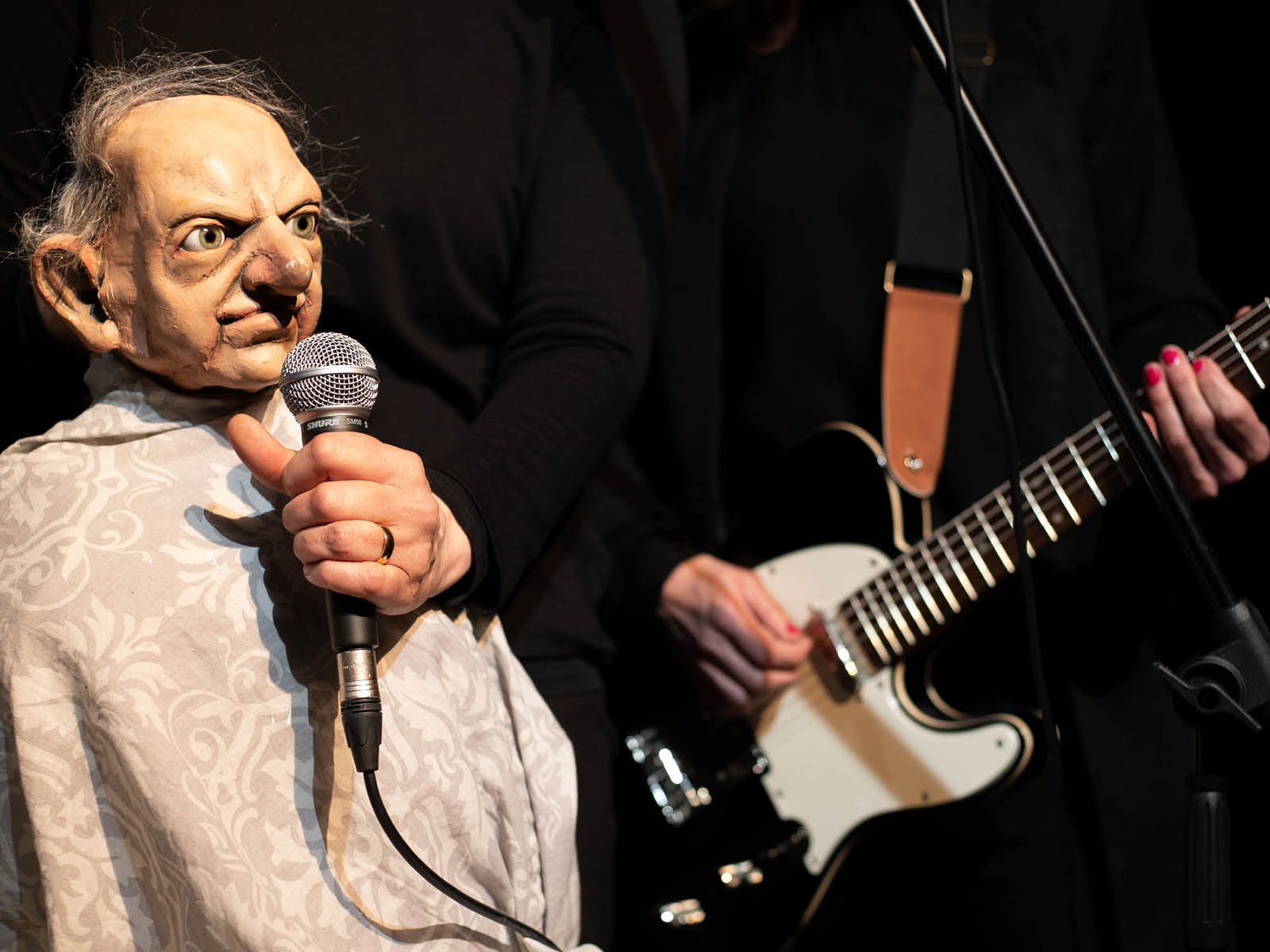The puppet of an old man with a half bald head and deep facial furrows holds a microphone in front of his face. A person plays an electric guitar in the background.
