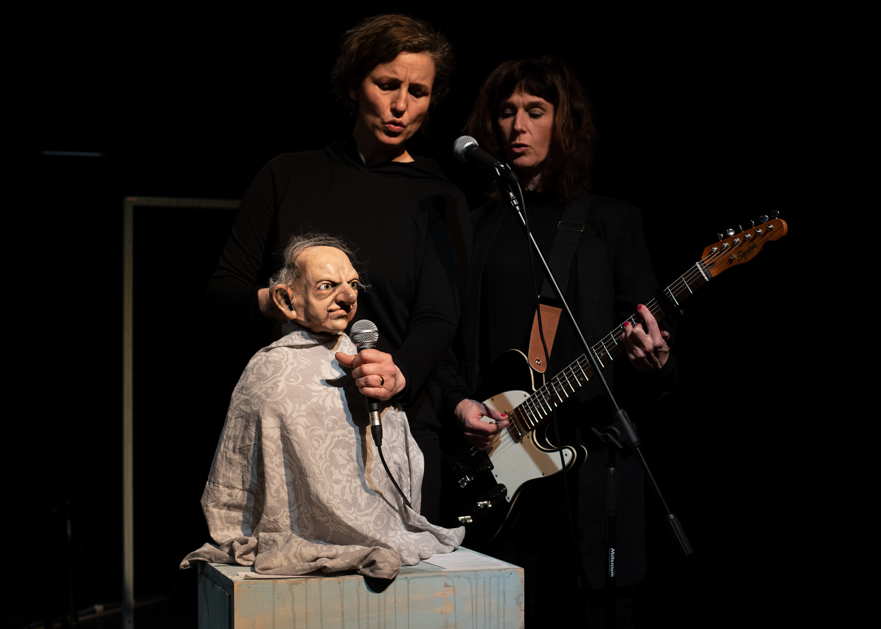 A puppet in the form of an old man sits on a small pedestal and holds a microphone in front of his face. In the background are two puppeteers dressed in black. One sings, the other plays the electric guitar.