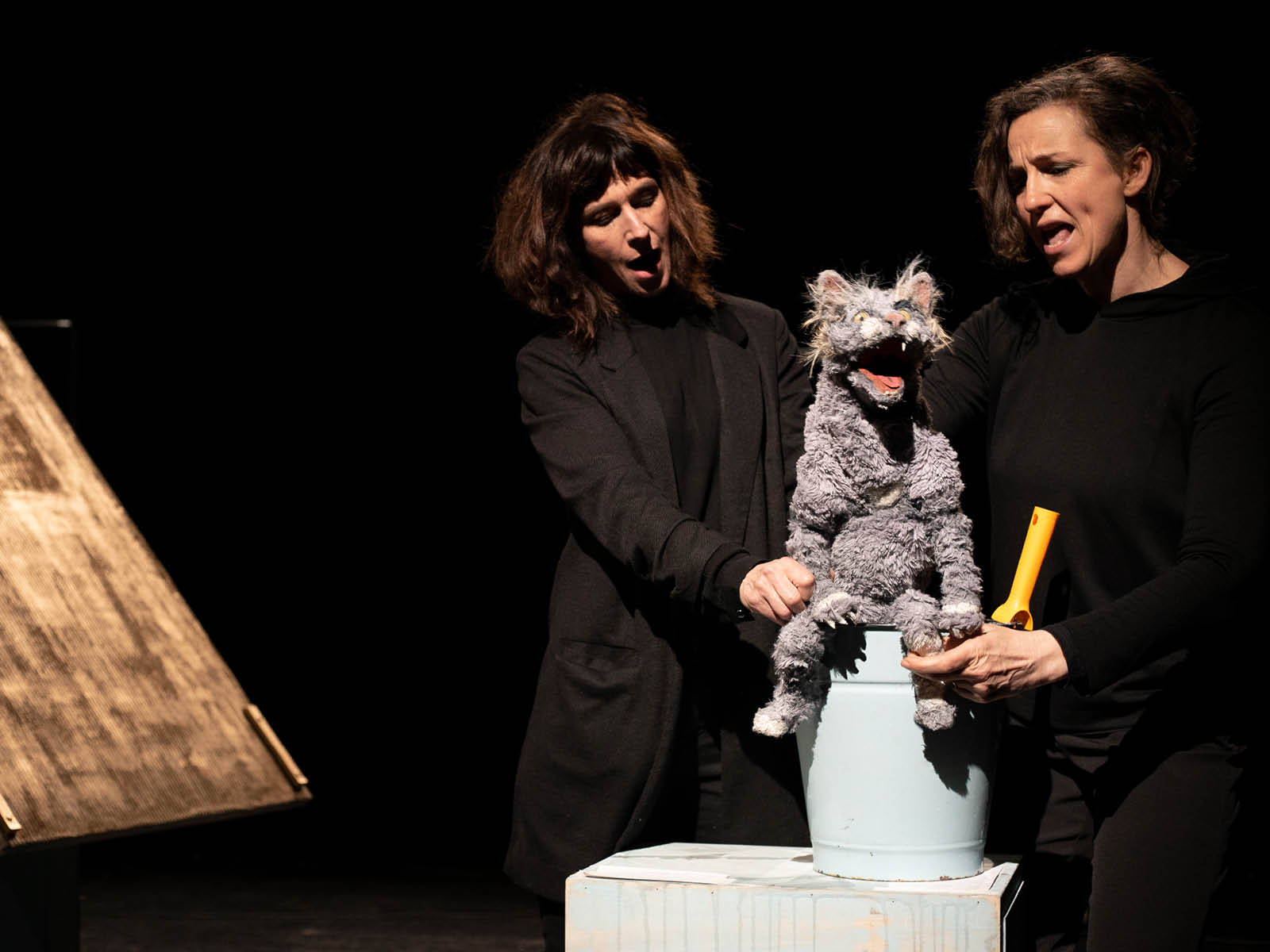 Two puppeteers dressed in black hold the puppet of a cat in their hands. The cat has its mouth wide open and is singing. It is sitting on an upturned metal bucket.