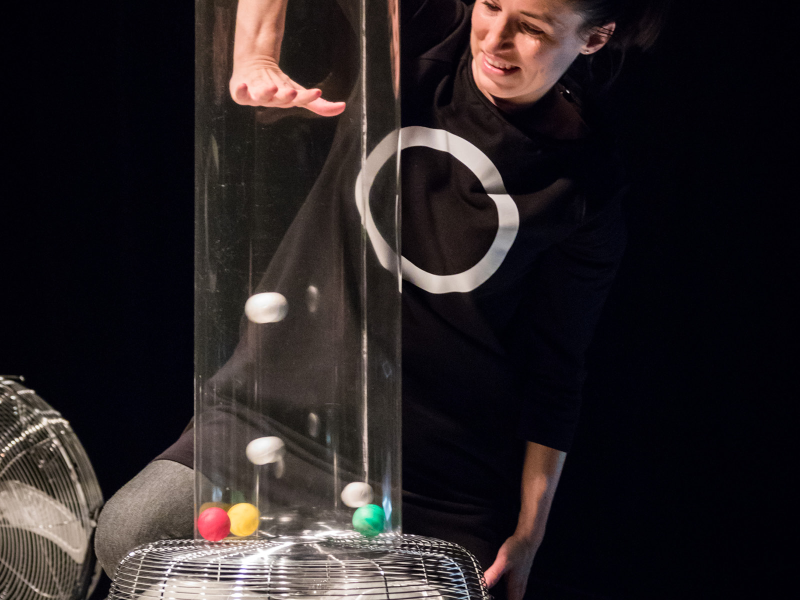 One of the performers regulates the trajectory of several small balls blown upwards by a fan by pushing her hand into the transparent tube in which the balls fly.