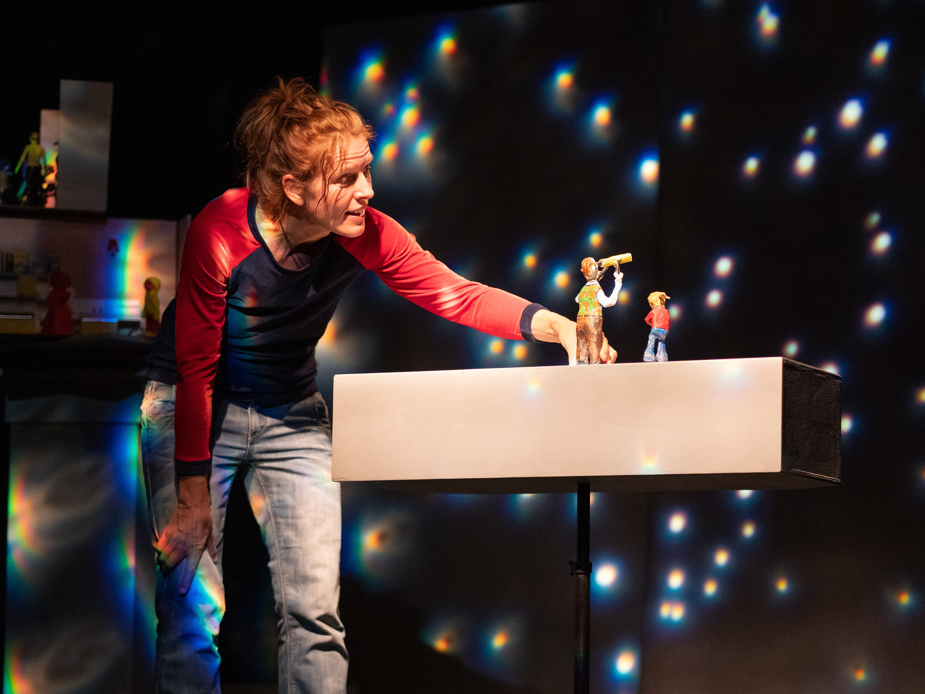 Puppenspielerin Annegret Geist beugt sich über ein längliches Brett in Hüfthöhe. Auf dem Brett steht die Figur eines kleinen Mädchens und eines Mannes mit einem Fernglas zum Sterne beobachten. Hinter den Beiden leuchten helle Punkte und simulieren einen Sternenhimmel.
