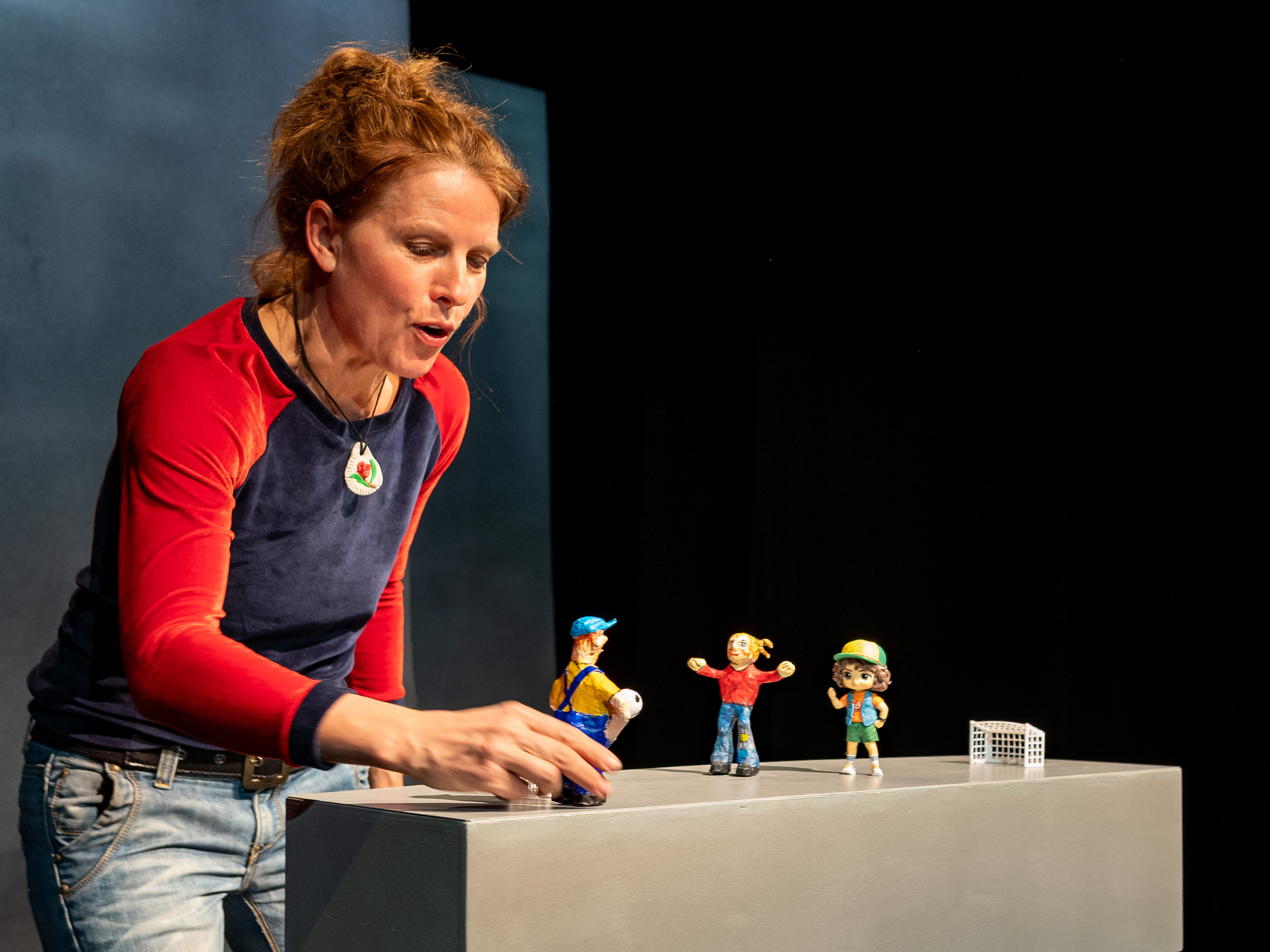 Puppenspielerin Annegret Geist hält die Figur eines Jungen in blauen Latzhosen und blauen Käppis in der Hand. Vor der Figur des Jungen steht die Figur eines kleinen Mädchens mit roten Haaren, daneben die Figur eines anderen Mädchens mit braunen Haaren und grüner Kappe.
