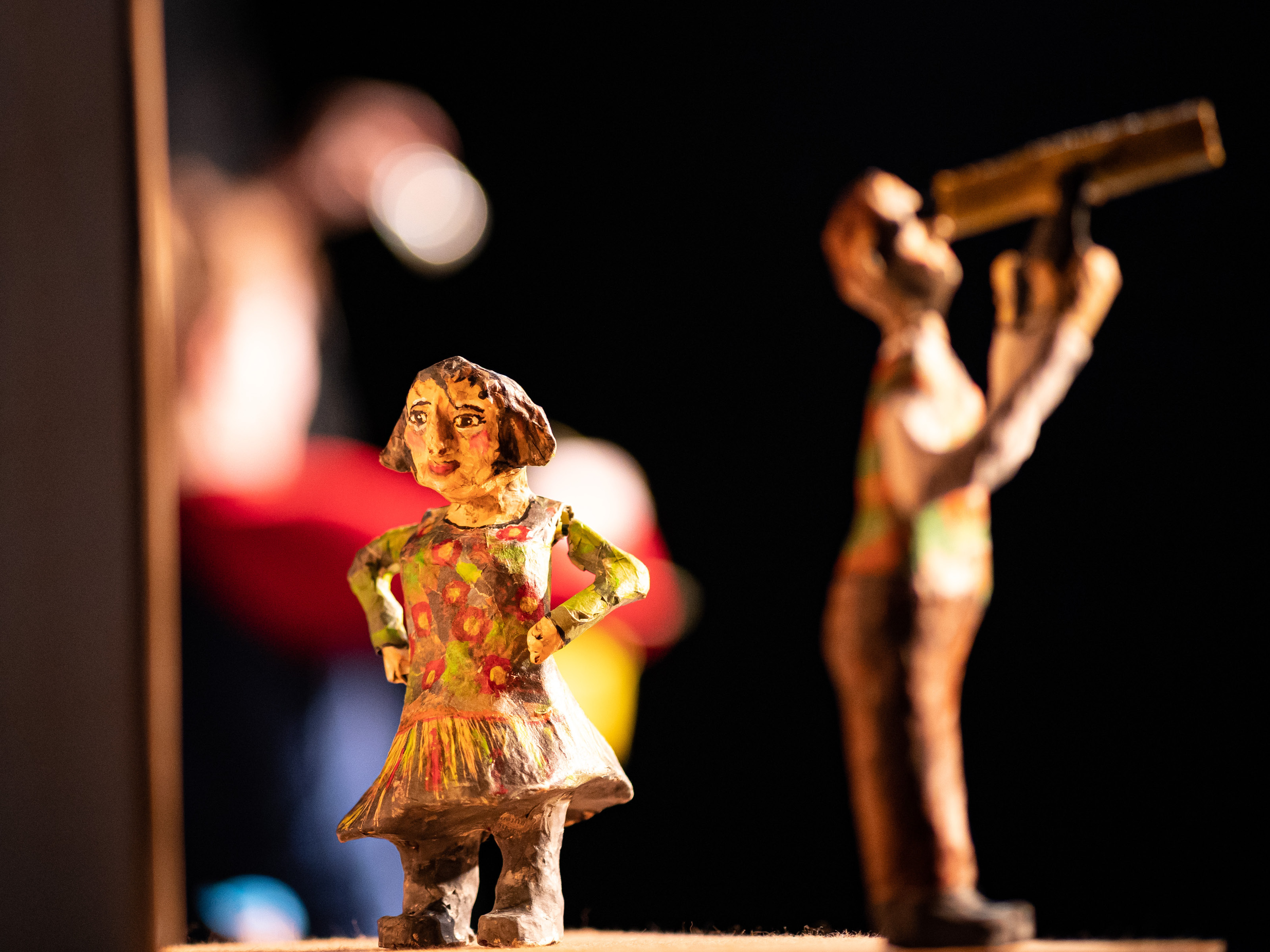 The clay figure of a little girl with a flowery dress and short brown hair looks past the viewer with her hands on her hips. In the background is a clay figure of a man with binoculars looking up at the sky.