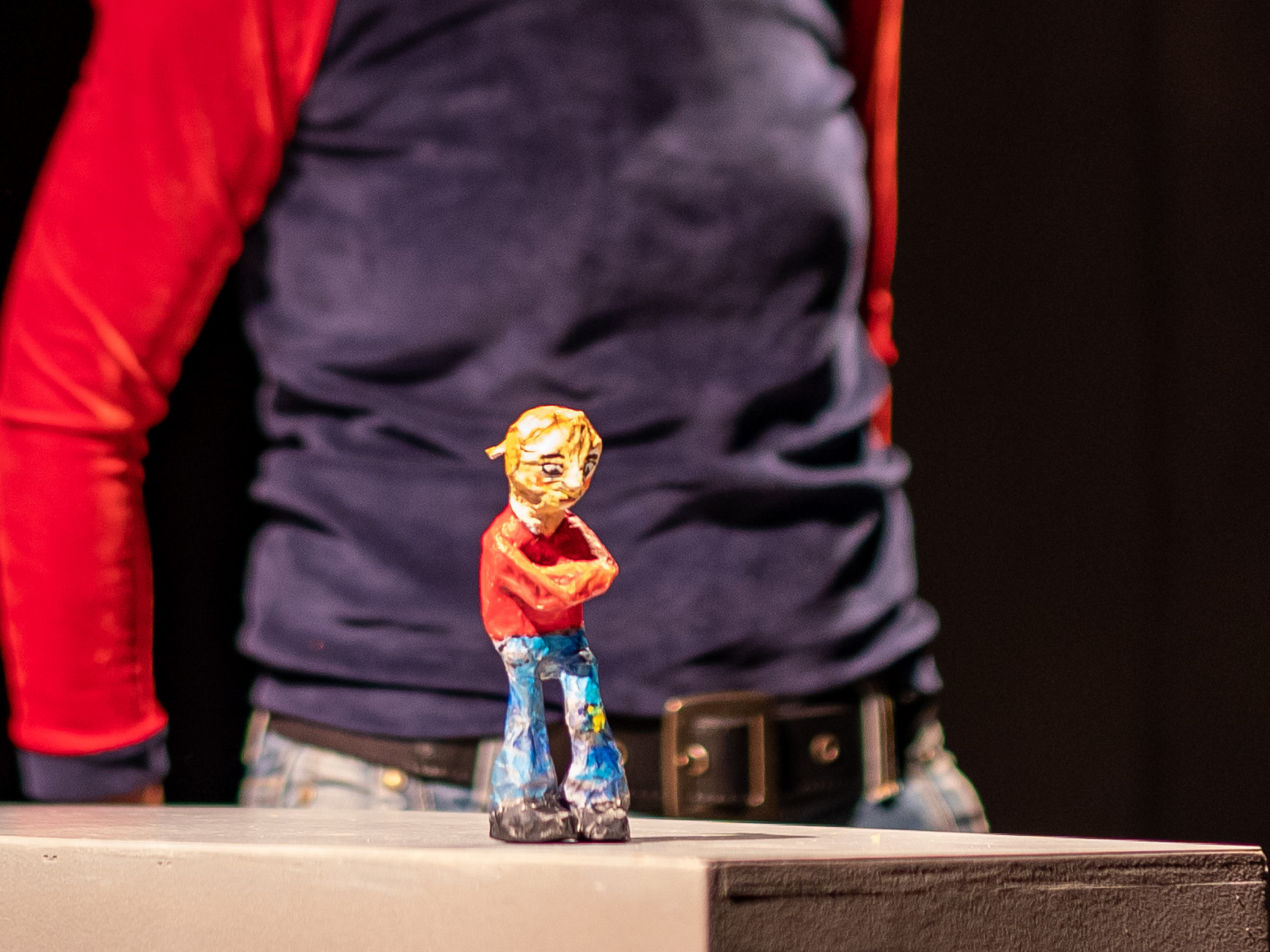 Die Figur eines kleinen Mädchens mit rotem Pullover, roten Haaren und blauen Schlaghosen steht mit verschränkten Armen und gesenktem Kopf im Vordergrund des Bildes. Im Hintergrund in Unschärfe ist der Oberkörper von Puppenspielerin Annegret Geist zu erkennen.