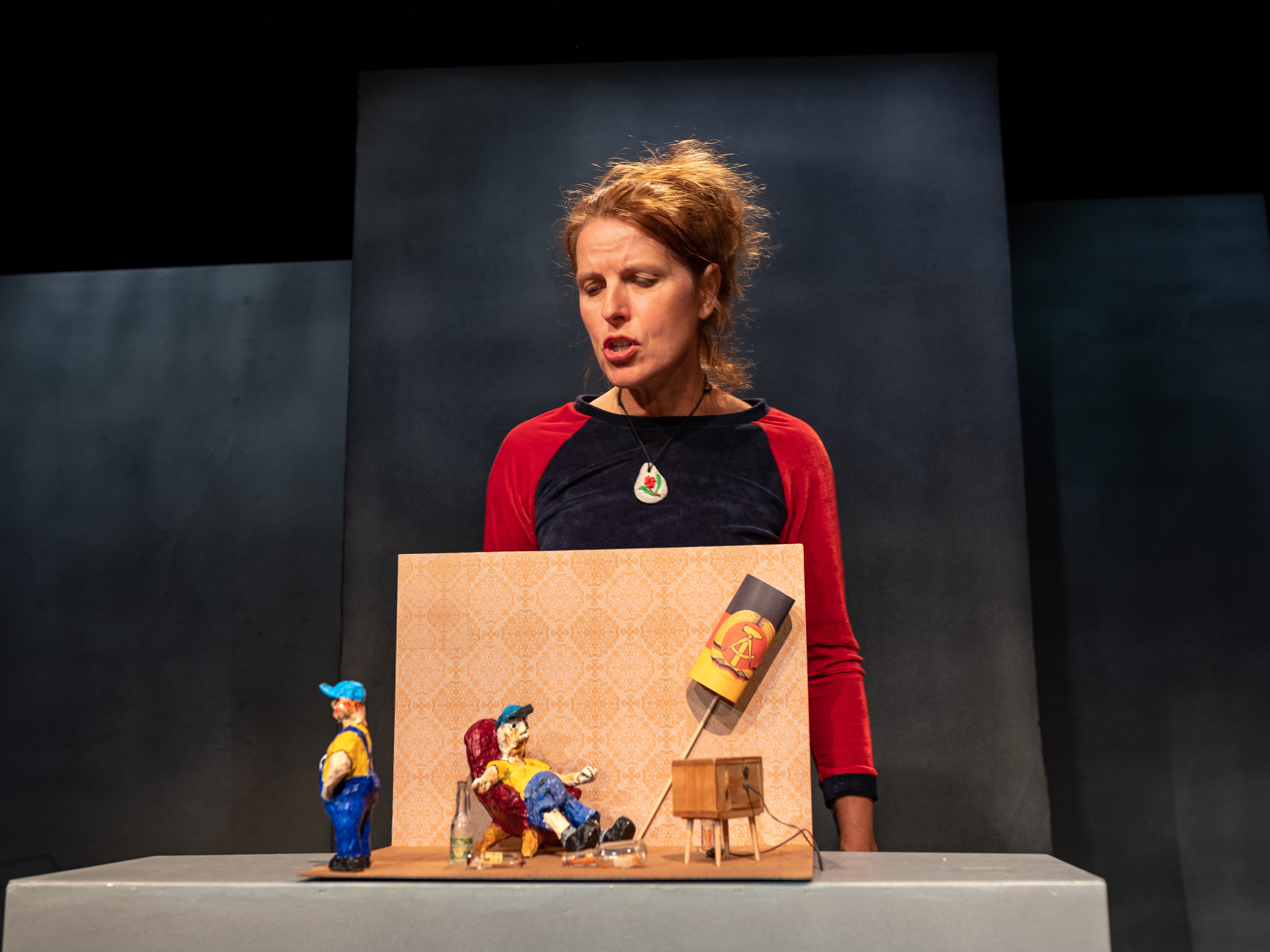 Puppeteer Annegret Geist stands behind the model of a living room. The figure of a man with a blue cap is sitting in the living room. He is watching television. Next to him is a lamp with a German flag. Turning away from the living room and the figure in the armchair is the figure of a little boy. He is also wearing a blue cap and blue pants.