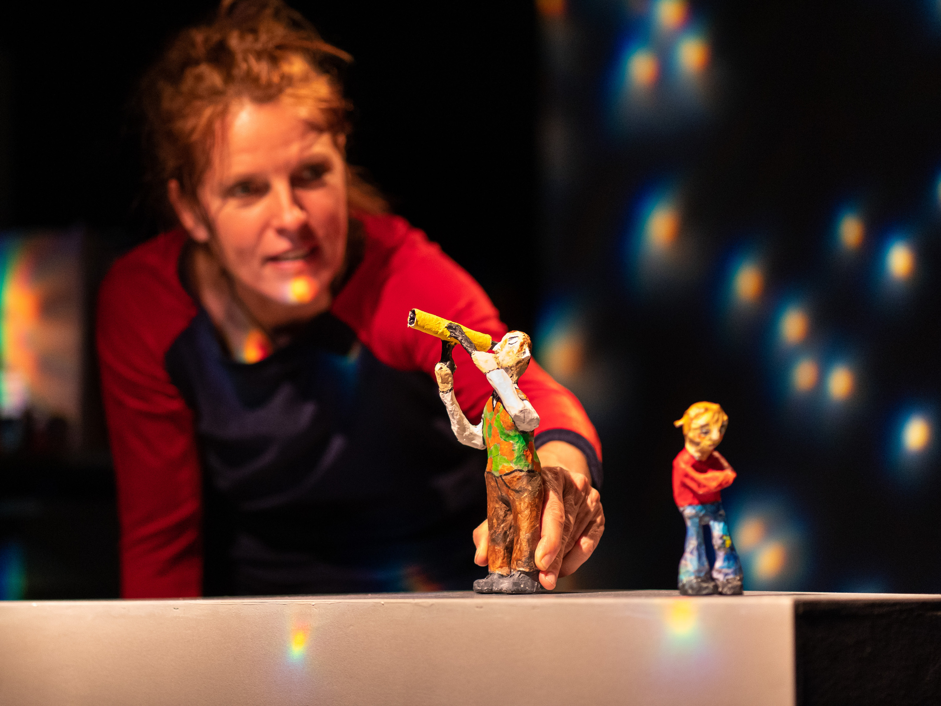 Puppeteer Annegret Geist holds the figure of a man with a telescope in his hand. A little girl with red hair stands facing away from him with her arms crossed. In the background, bright points of light shine on the dark wall.