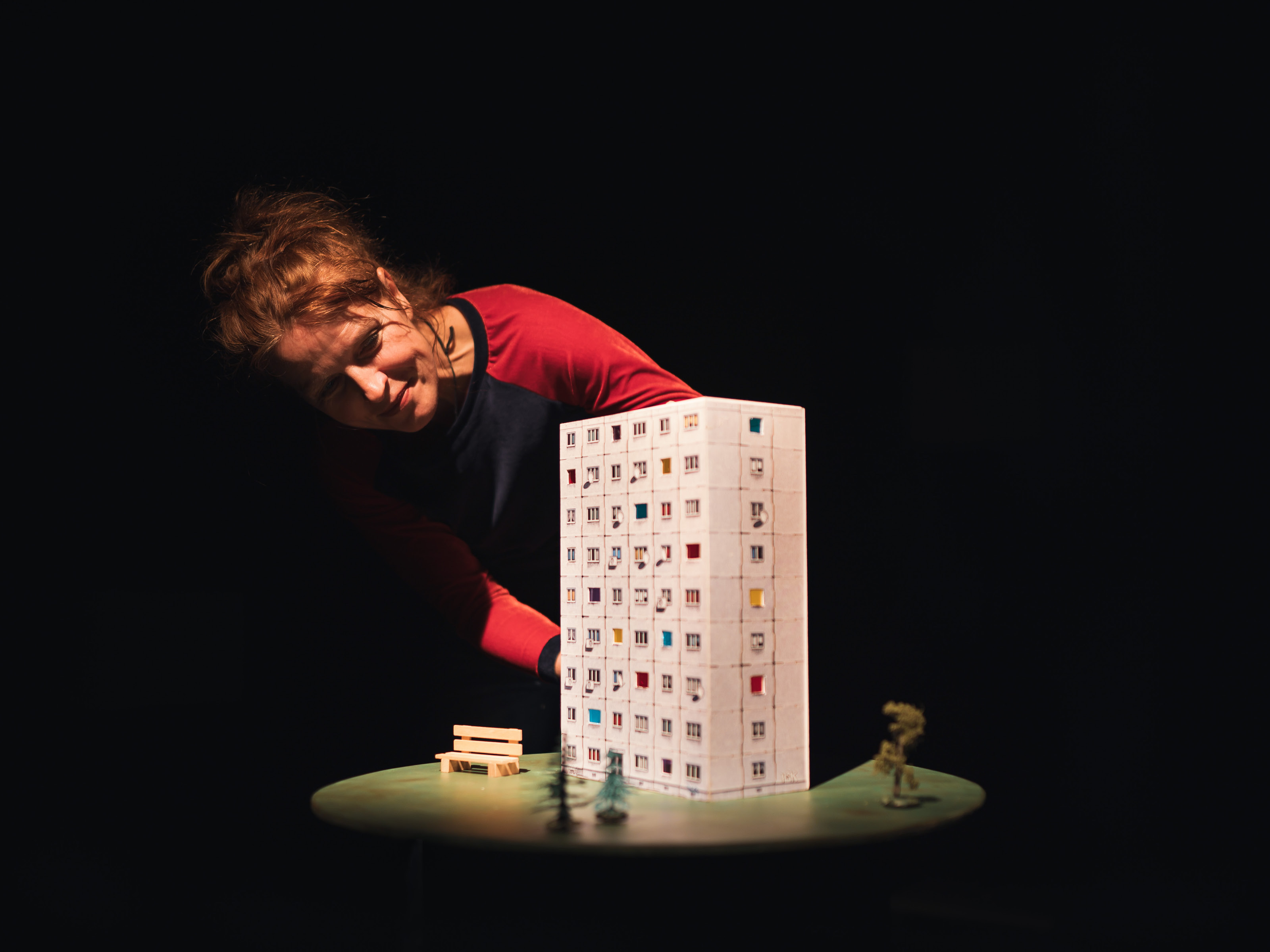 Puppenspielerin Annegret Geist beugt sich über ein Hochhaus mit neun Stockwerken. Vor dem Hochhaus steht eine kleine Holzbank auf einer angedeuteten Grünfläche.
