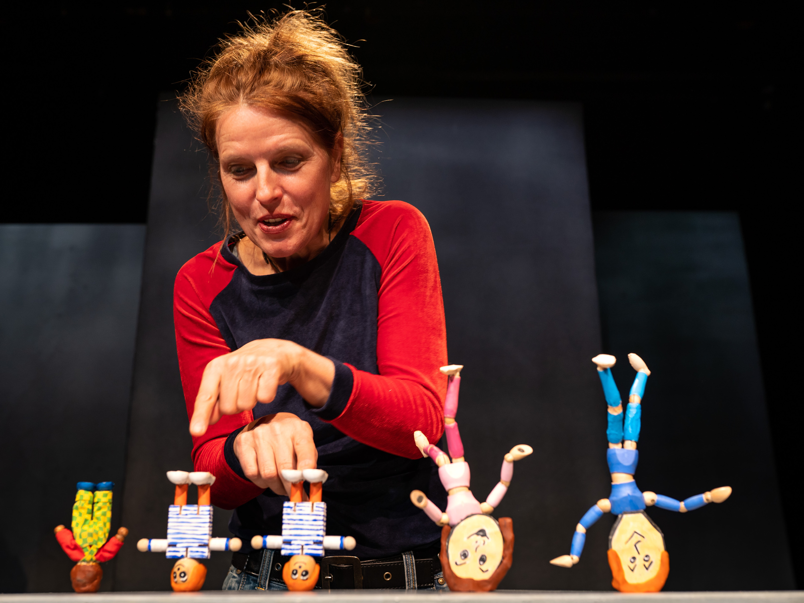 Five figures stand upside down in front of puppeteer Annegret Geist, their legs stretched up in the air. She has stretched out her finger and is pointing at one of the smaller figures.