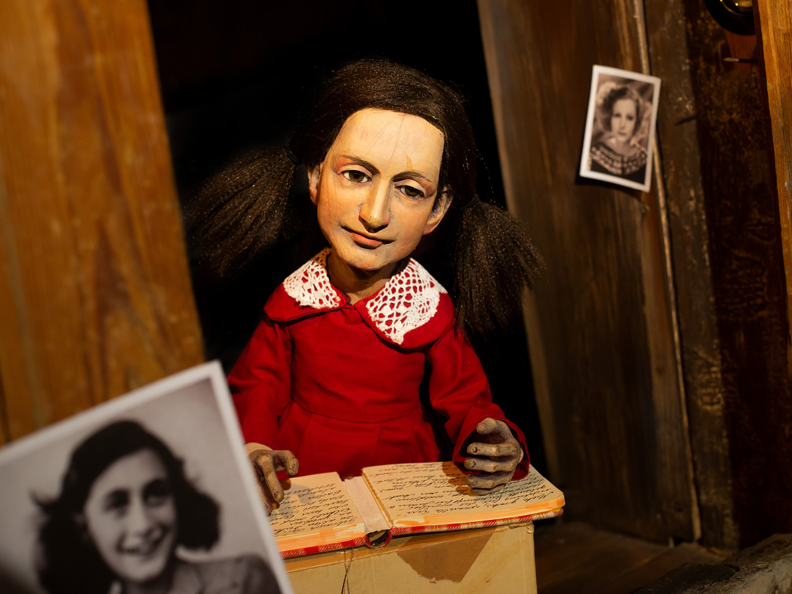 In rotem Kleid, mit Spitzenkragen und zwei Zöpfen rechts und links, sitzt die aus Holz gefertigte Puppe von Anne Frank an einem Tisch. Ihr Tagebuch ist offen vor ihr ausgebreitet. Im Vordergrund ist unscharf eine Fotografie der echten Anne Frank zu sehen.