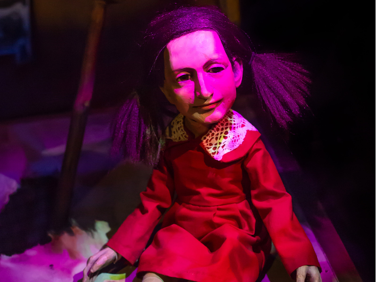 Die Puppe Anne Frank sitzt auf dem Boden und wird von magentafarbenem Licht angestrahlt. Ihr Gesicht ist nach rechts gewandt, die Gesichtszüge sind untergründlich aber die Mundwinkel zeigen ein wenig nach oben.