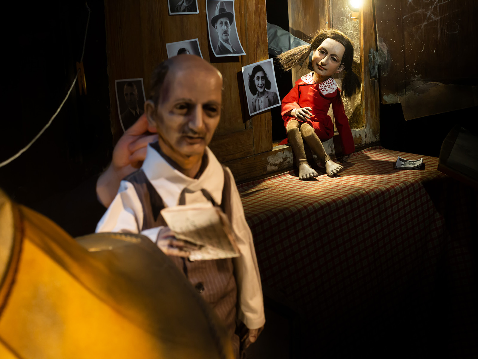 Die Puppe eines Mannes mit Halbglatze, tiefen Schatten unter den Augen und eleganten Klamotten hält einen gefalteten Brief in der Hand. Im Hintergrund ist die barfüßige Puppe von Anne Frank, umgeben von Fotografien zu erkennen.