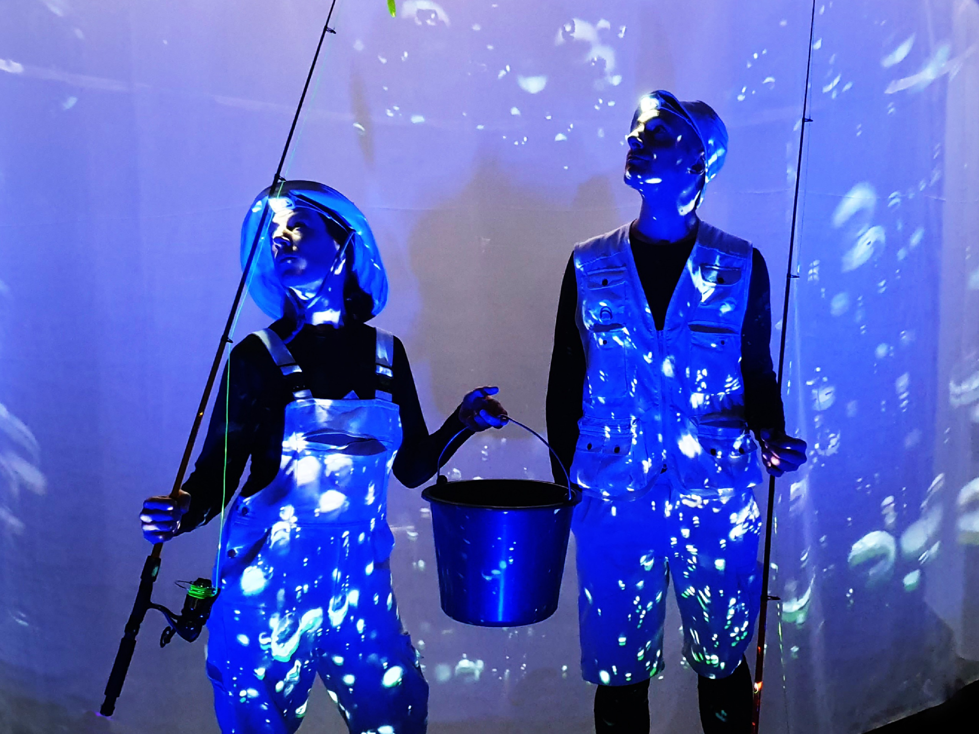 Two people in fishing outfits stand in front of a blue lit screen with bubbles and hold two fishing rods and a metal bucket in their hands.