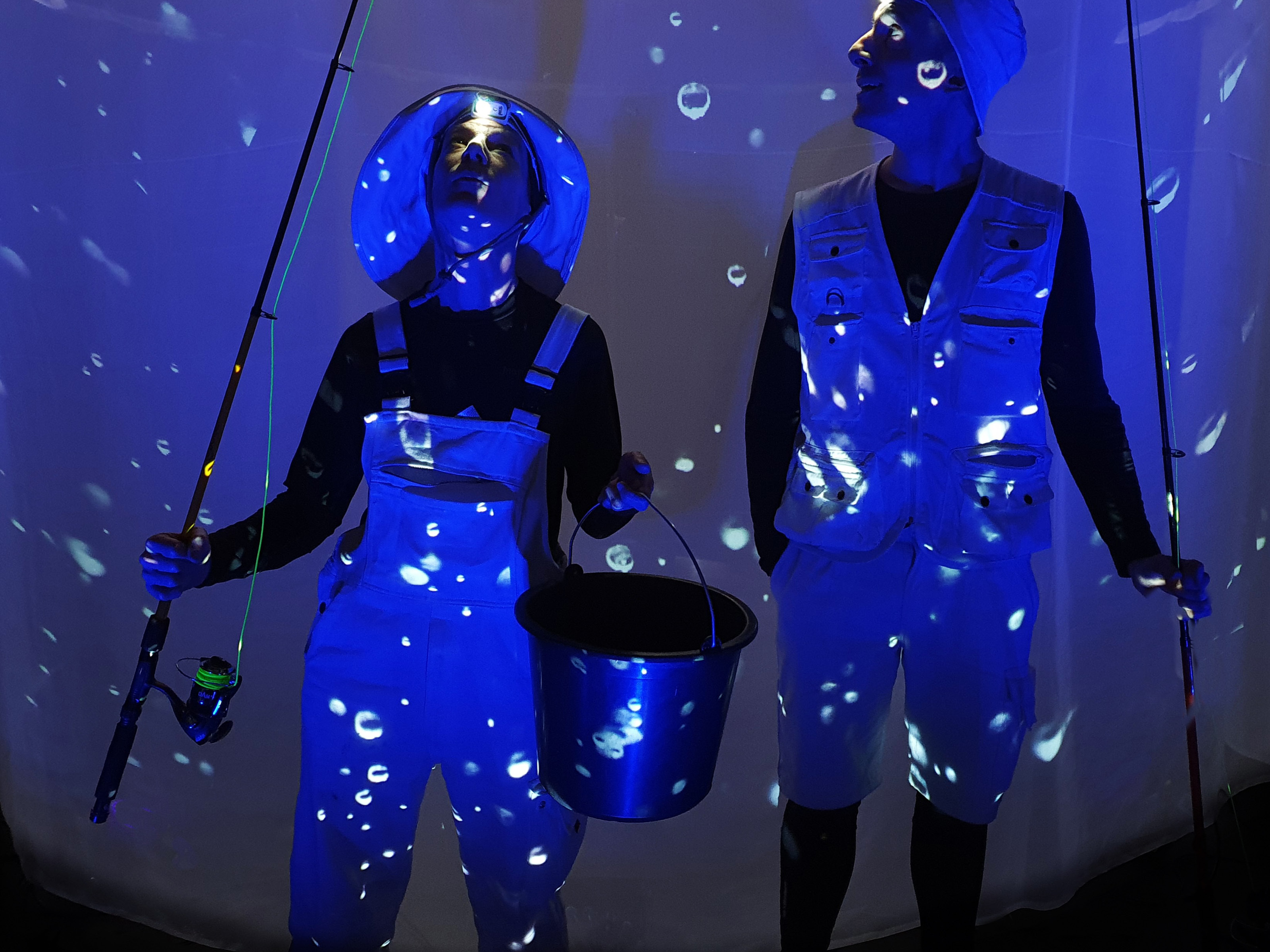 Two people in fishing outfits stand in front of a blue lit screen with bubbles and hold two fishing rods and a metal bucket in their hands.