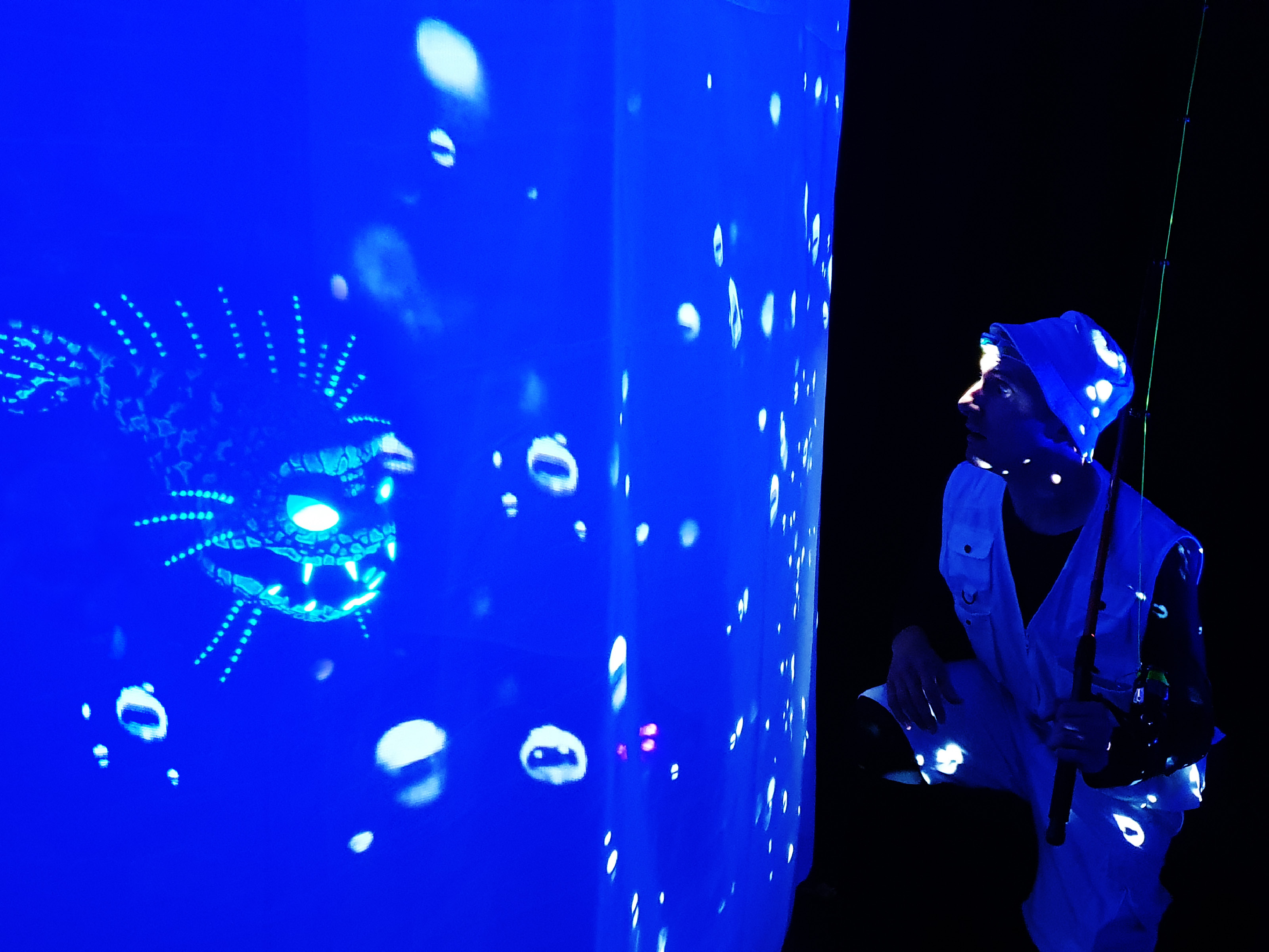 On a white projection surface, a fish with jags and pointed teeth is projected onto a blue background. In front of the fish projection kneels a man with fishing vest, trousers, hat and fishing rod and looks at him.