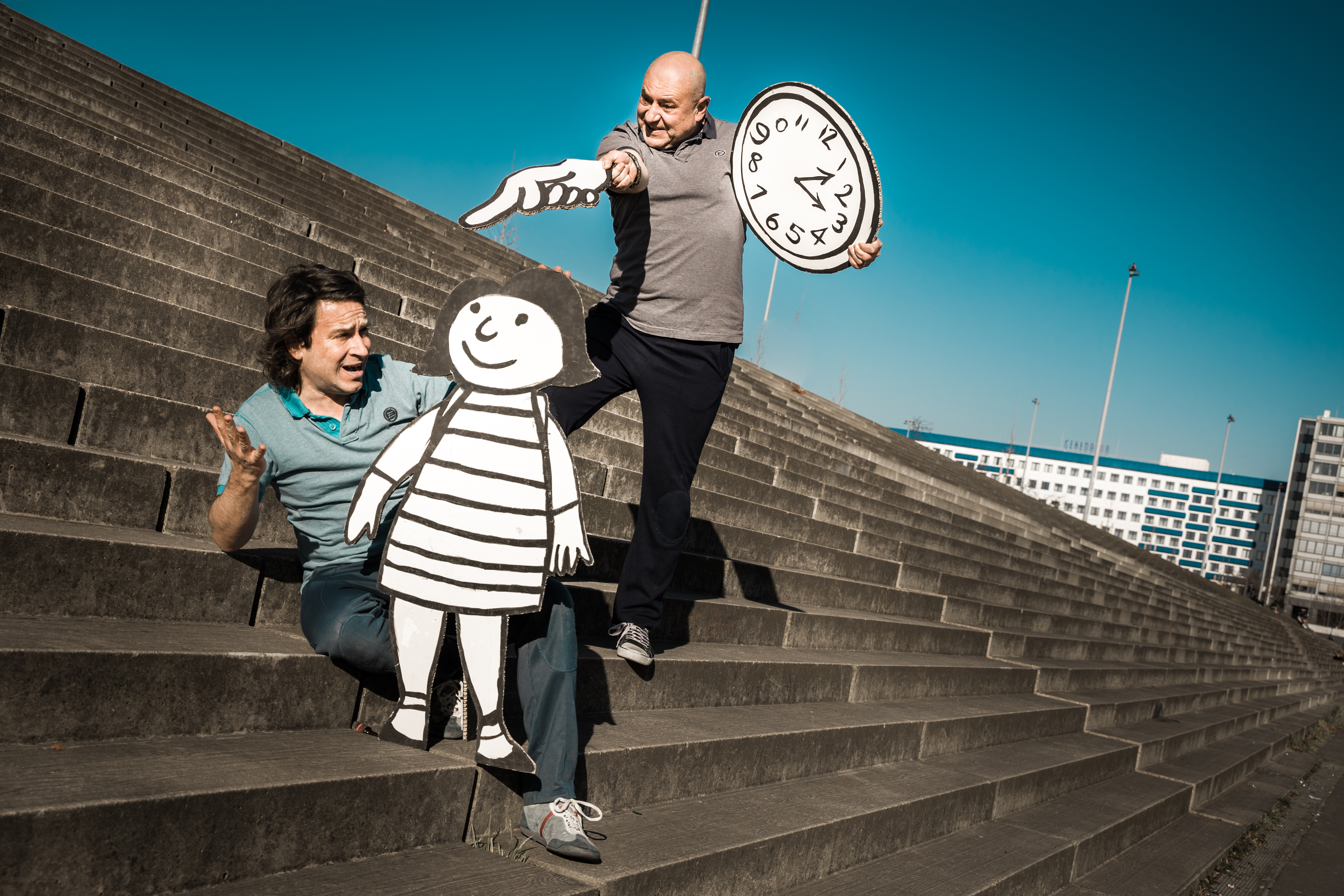 Zwei Spieler mit einer lebensgroßen Flachfigur und einer Uhr aus Papier auf Treppen posierend.