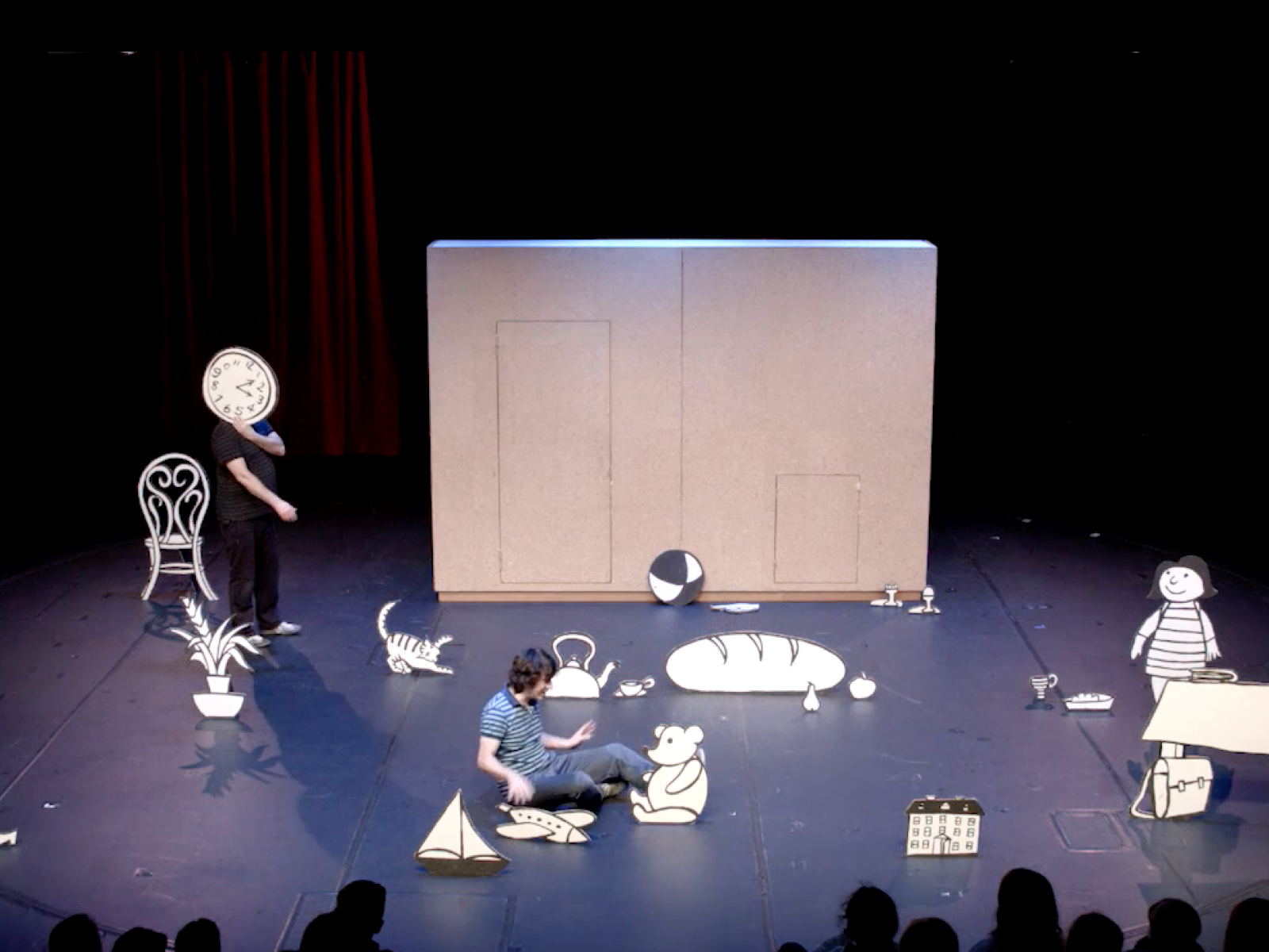 Bühne mit weißer Hintergrundwand. Im Vordergrund zwei Puppenspieler mit bemalten Figuren aus Pappe. Weitere Objekte sind auf dem Bühnenboden verteilt.