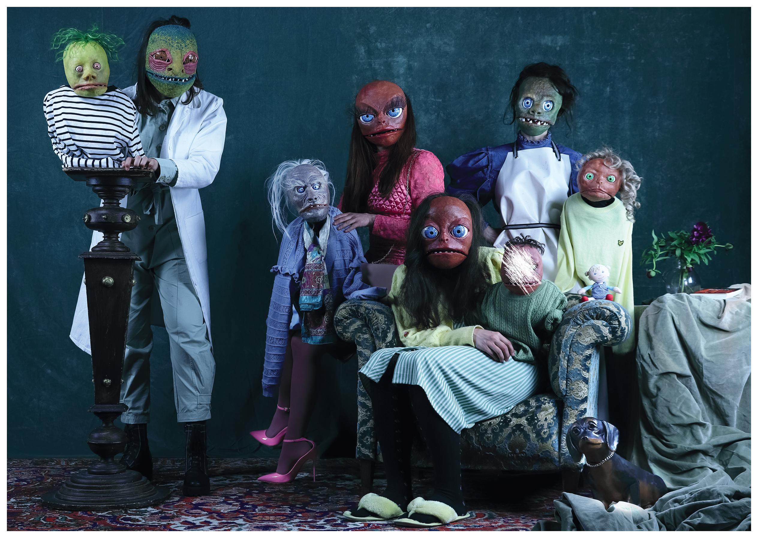 Gruppenbild der Figuren von Creatures Hill: Vier Schauspielerinnen tragen traurig bis grotesk wirkende Masken, die etwas an Reptilien erinnern, und je eine Klappmaulpuppe auf dem Arm. Auf dem Bild ist das Gesicht einer der Puppen weggekratzt.