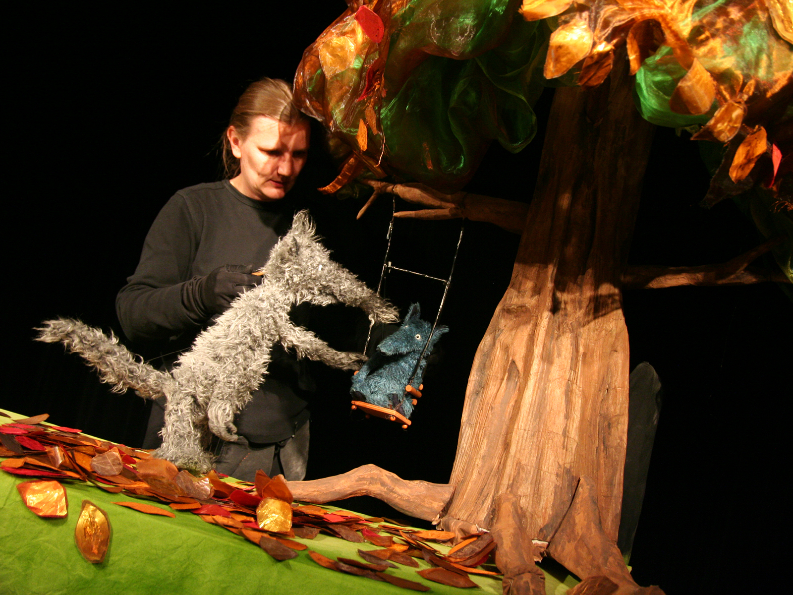 Der große Wolf schubst eine Schaukel an, in dem der kleine Wolf sitzt. Die Schaukel hängt an einem Baum. Hinter den Figuren ist die Puppenspielerin abgebildet.
