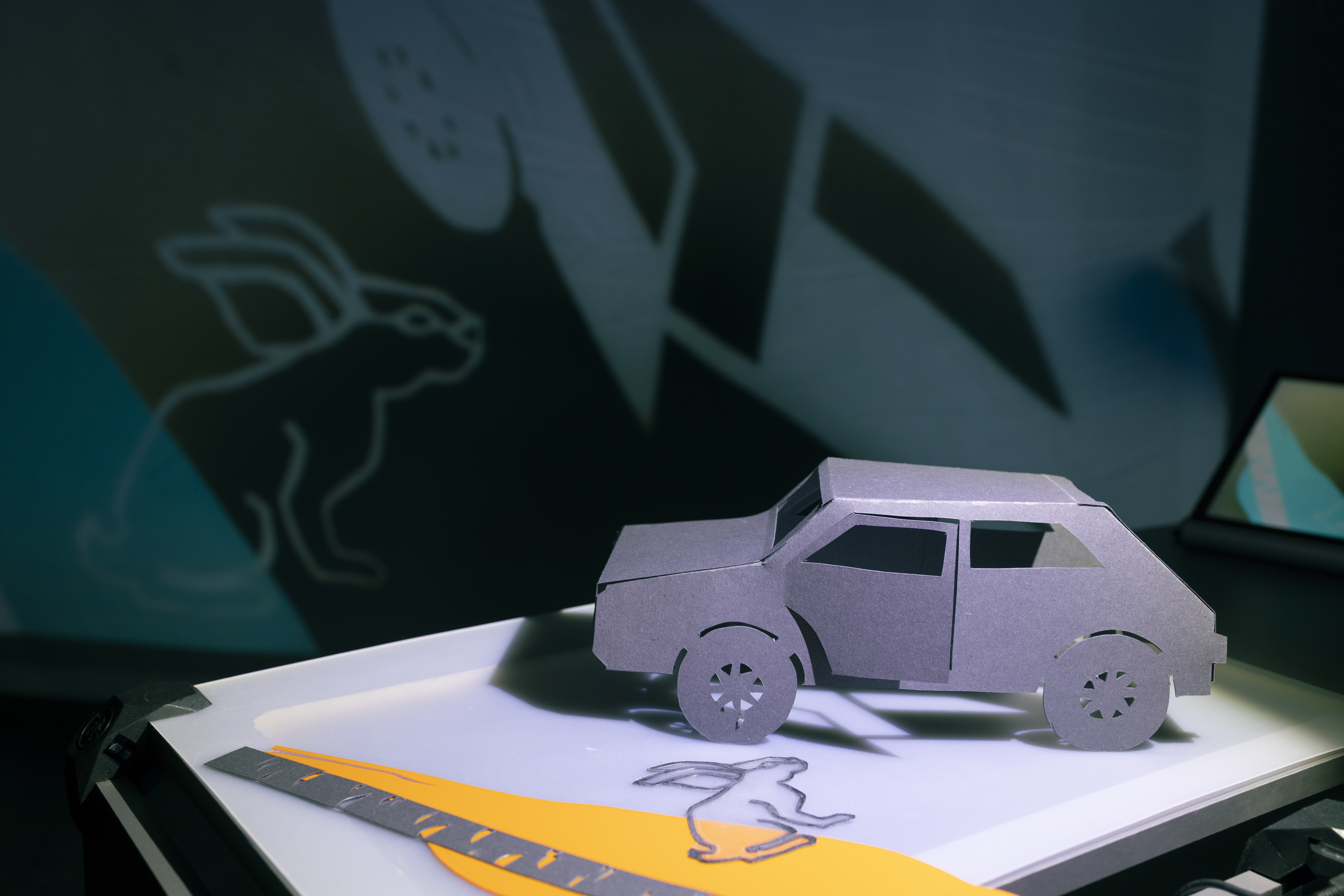 Ein Auto und ein Hase aus Papier auf einer weißen eckigen Fläche. An der Wand dahinter sind Projektionen ihrer Schatten abgebildet.