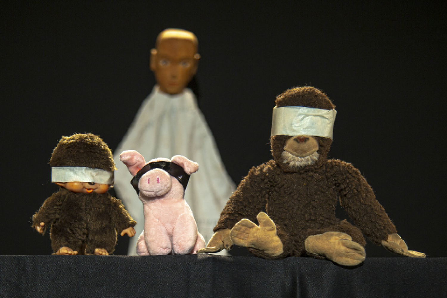 Drei Kuscheltiere mit verbundenen Augen: zwei Affen und ein kleines Schweinchen. Hinter ihnen steht eine Puppe mit weißem Gewand und hölzernem Glatzkopf.