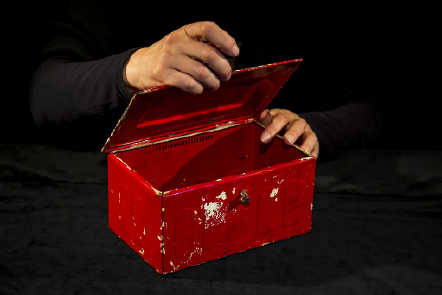 Dunkel gekleideter Performer öffnet eine rote Kiste. Von ihm sind nur die Hände zu sehen.