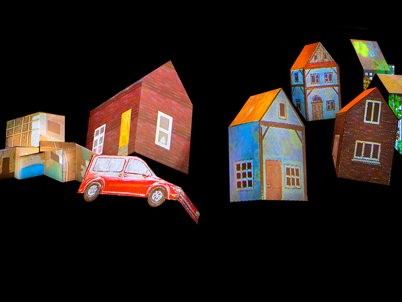 Häuser und Autos aus angemalter Pappe vor schwarzem Grund.
