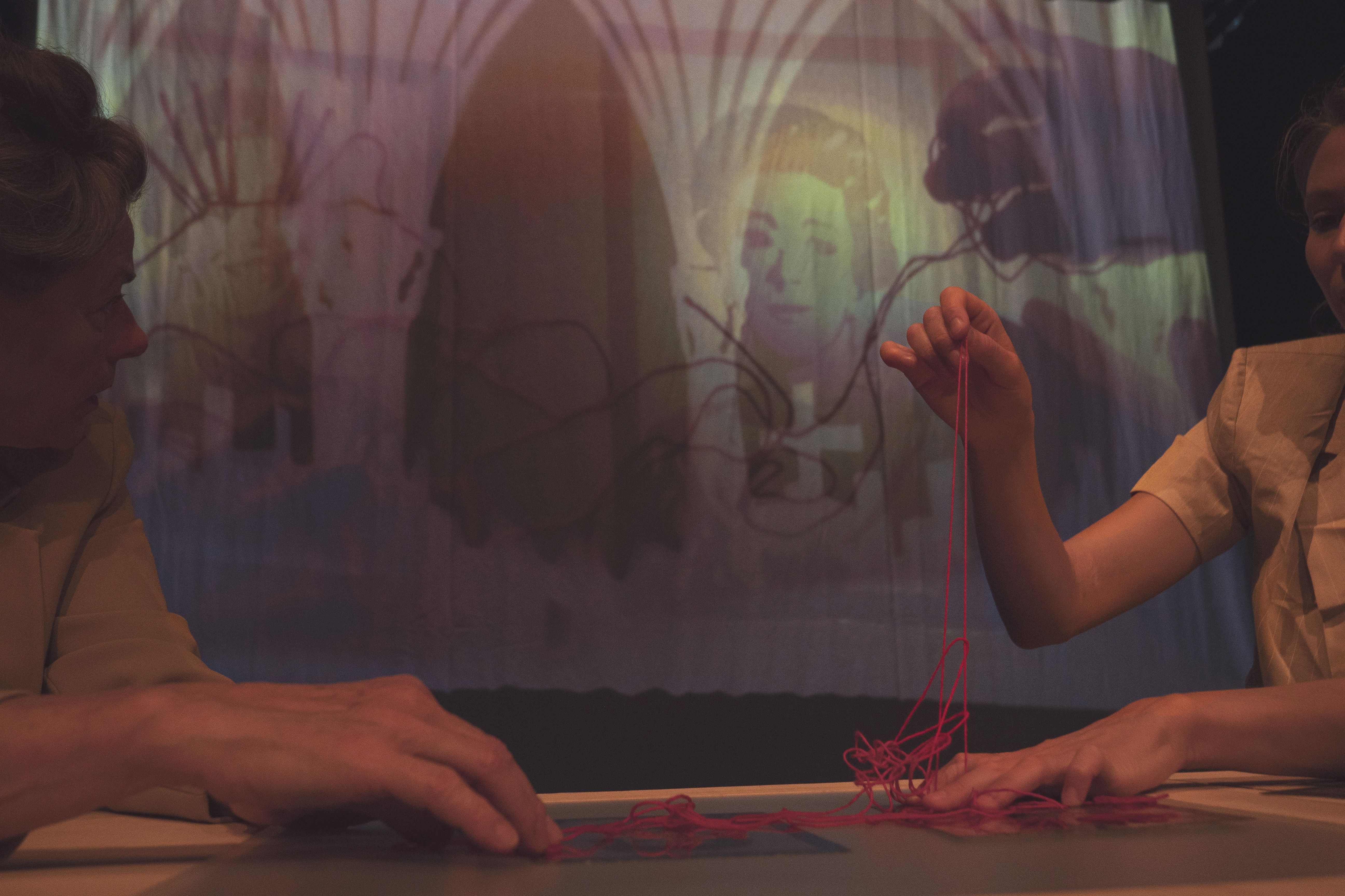 Vor einer Videoprojektionsfläche sind die Hände von zwei Schauspielerinnen im Bild, die einen roten Faden aus einem Knäuel ziehen.