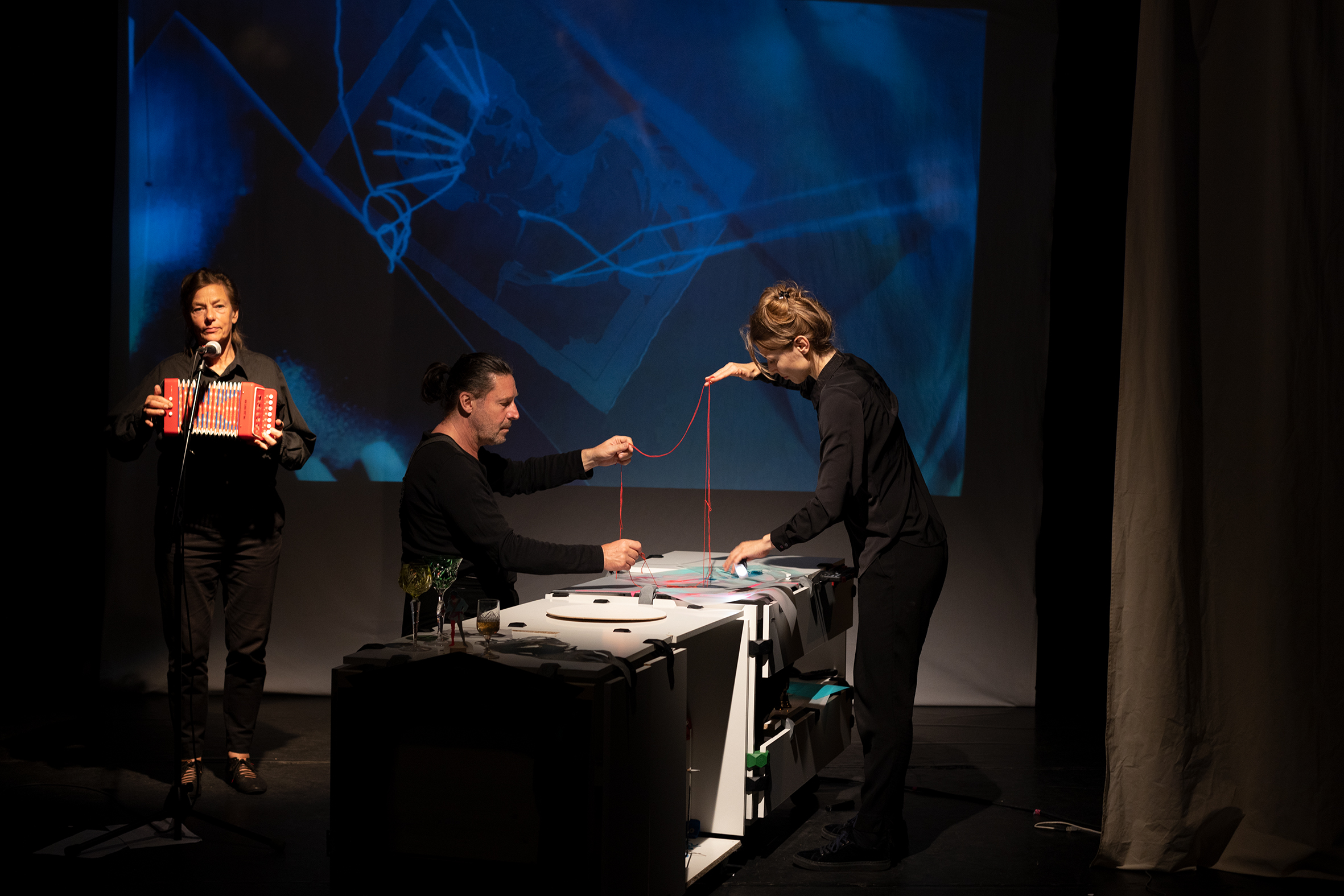 Zwei Schauspieler an einem Tisch mit Dingen aus Papier halten zusammen einen roten Faden. Links neben ihnen steht eine weitere Person auf der Bühne mit einem roten Kinder-Akkordeon. Im Hintergrund ist auf einer Leinwand eine Video-Projektion zu sehen.